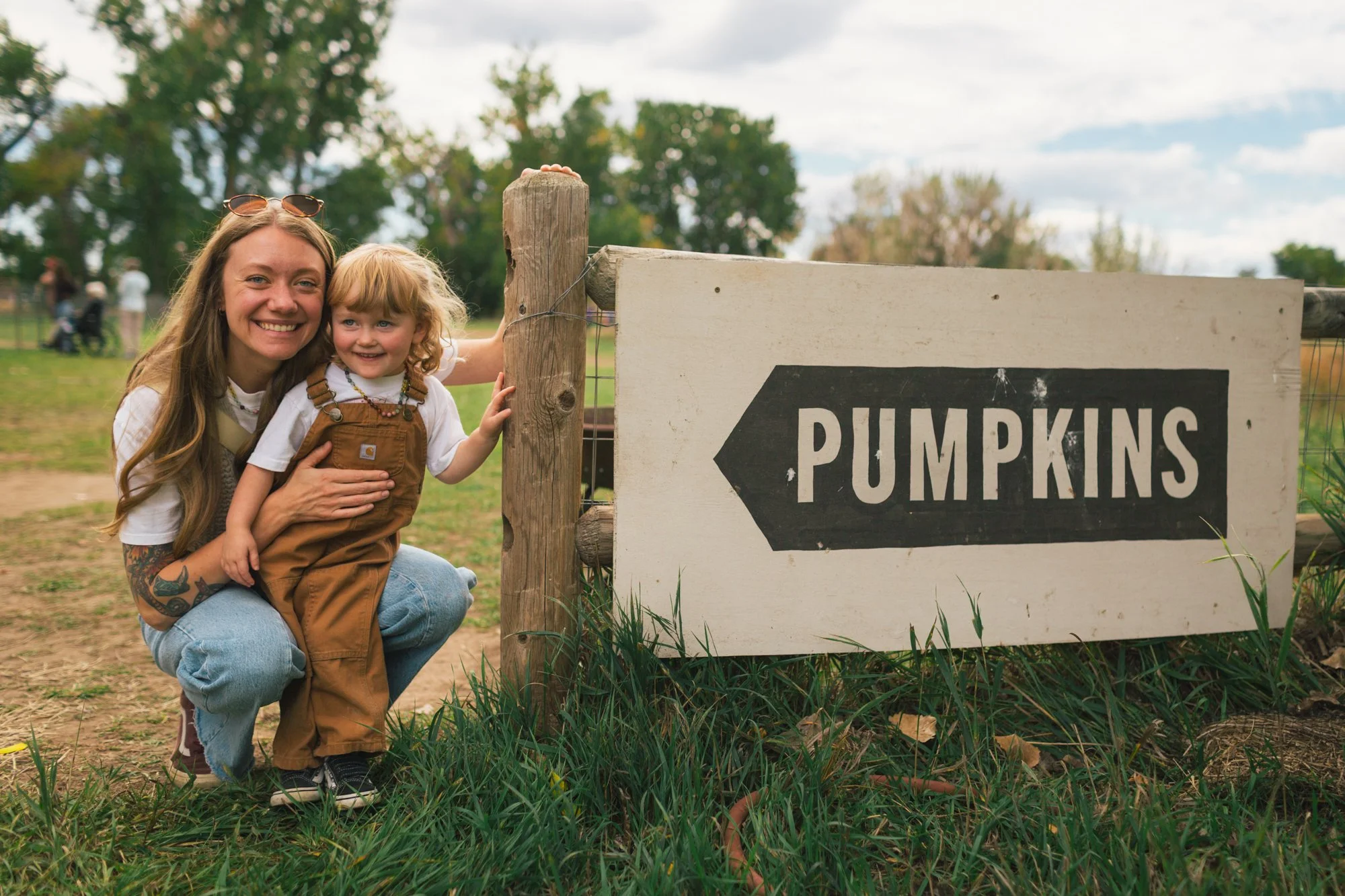 A woman with long hair, sunglasses on her head, and tattoos is crouching next to a young girl with curly blonde hair, wearing a brown apron over white t-shirt, in front of a wooden sign that reads 'PUMPKINS' with an arrow pointing to the right. They 