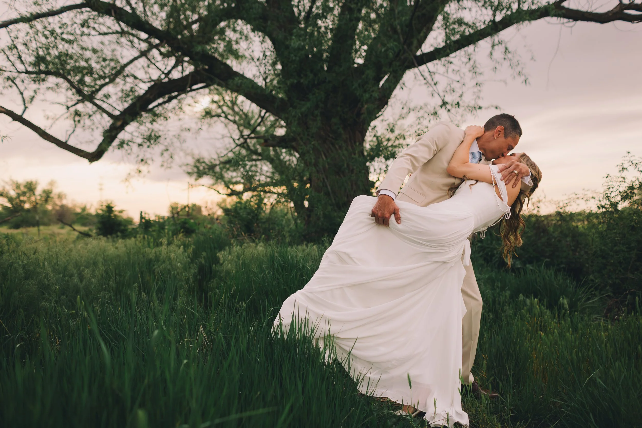 A couple sharing a kiss outdoors under a large tree, with the man holding the woman in his arms, dressed in wedding attire, during sunset or dusk.