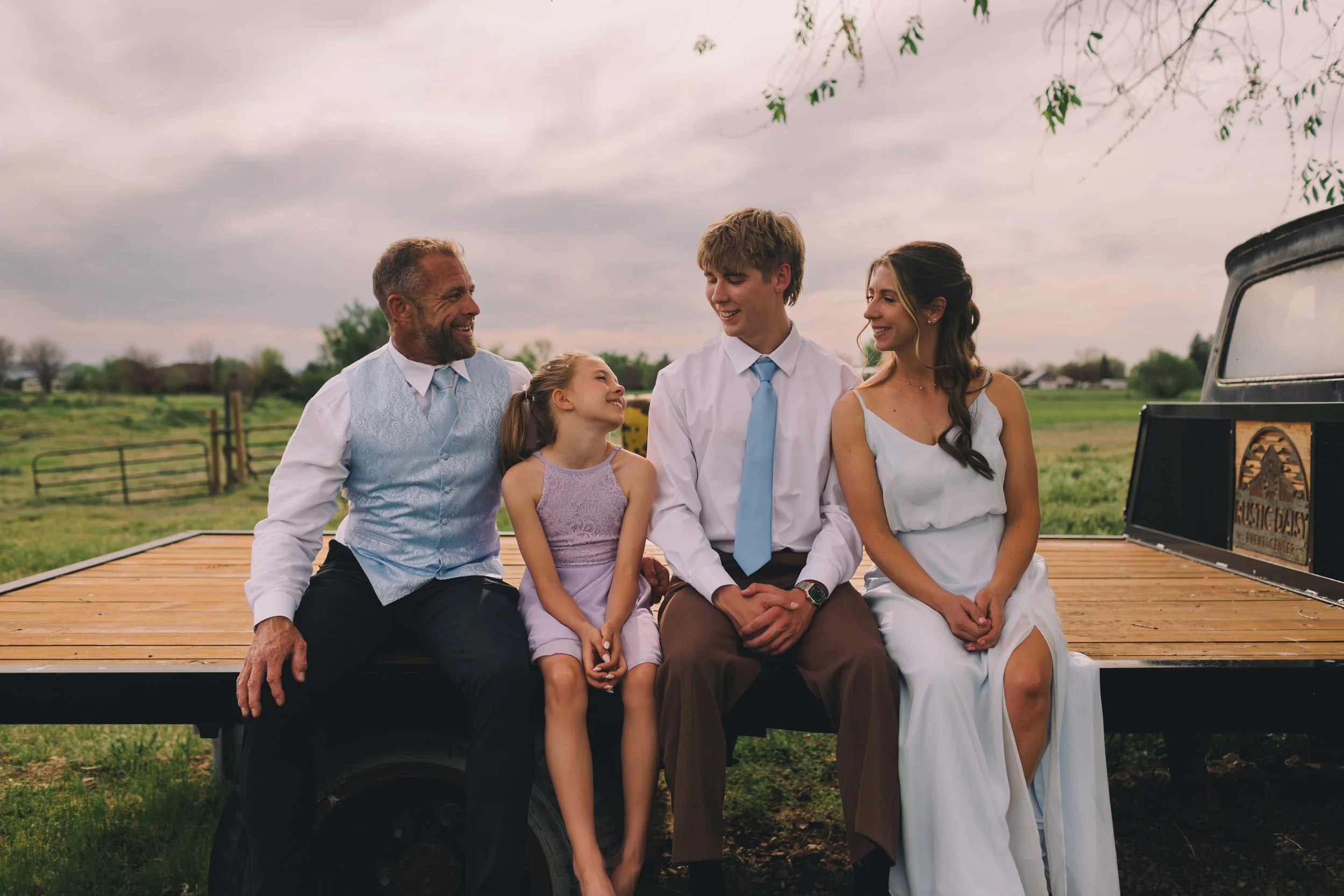 A family of five sitting together on a wooden trailer in an outdoor rural setting, smiling and enjoying each other's company during a cloudy day.