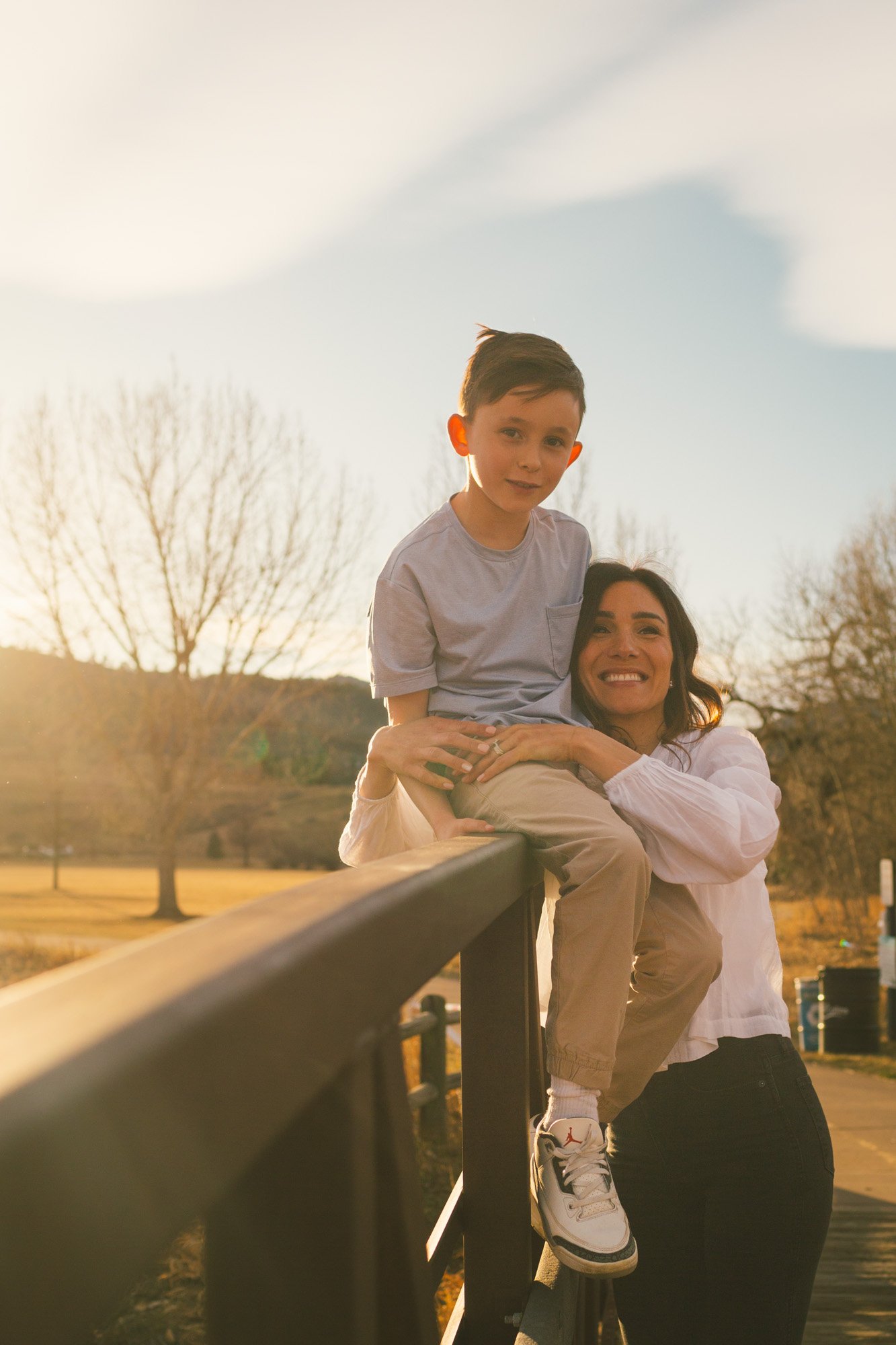 A woman lifting a young boy onto a bridge railing outdoors during sunset, with leafless trees and open field in the background.