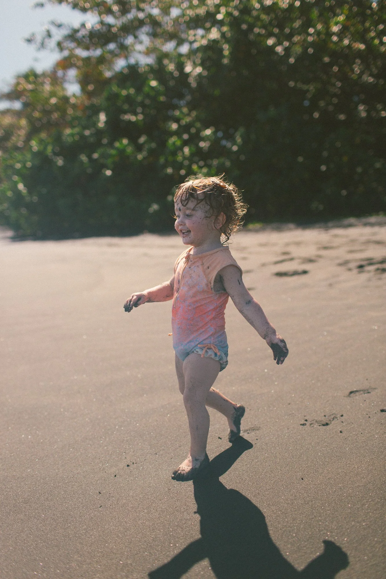 A young girl running on the beach, covered in sand and mud, wearing a sleeveless orange shirt and shorts, with a joyful expression.
