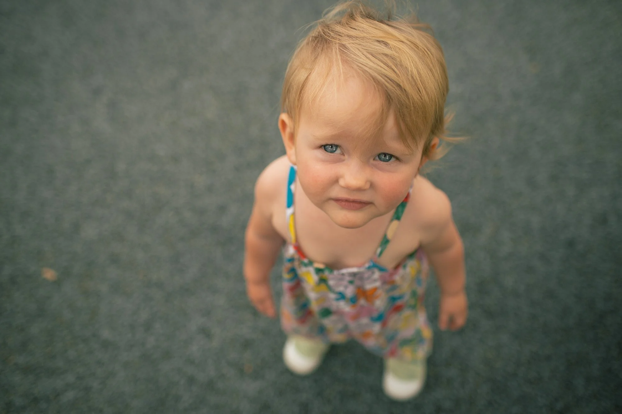 Young child with red hair looking up at the camera, wearing a colorful dress, on a gray outdoor surface.