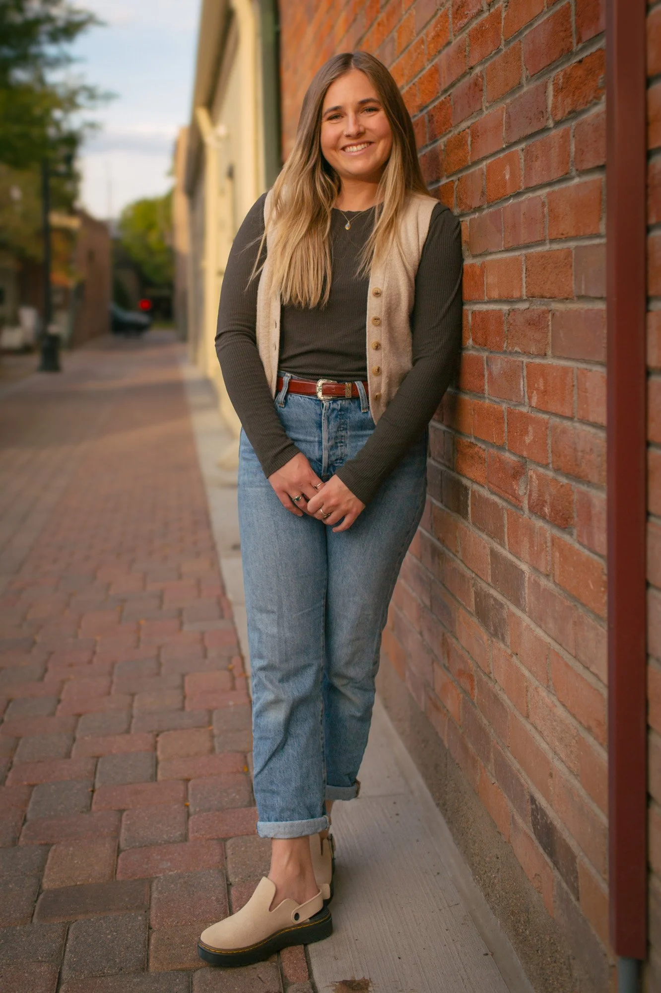 A young woman with long blonde hair leaning against a brick wall on a city sidewalk, smiling at the camera, wearing a black long-sleeve top, beige vest, jeans, and beige shoes.