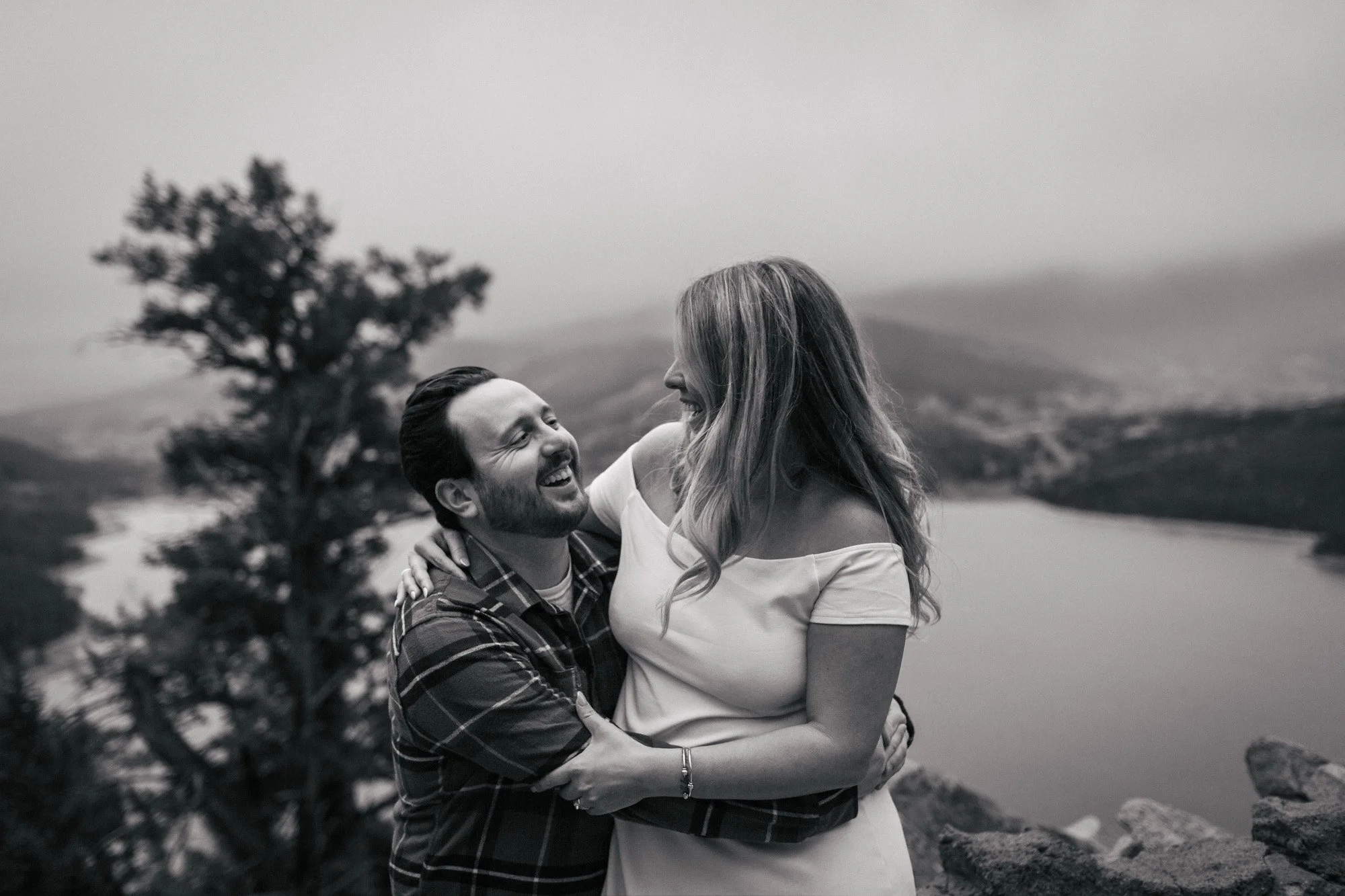 A black and white photo of a smiling couple holding each other outdoors on a rocky shoreline with hills and trees in the background.