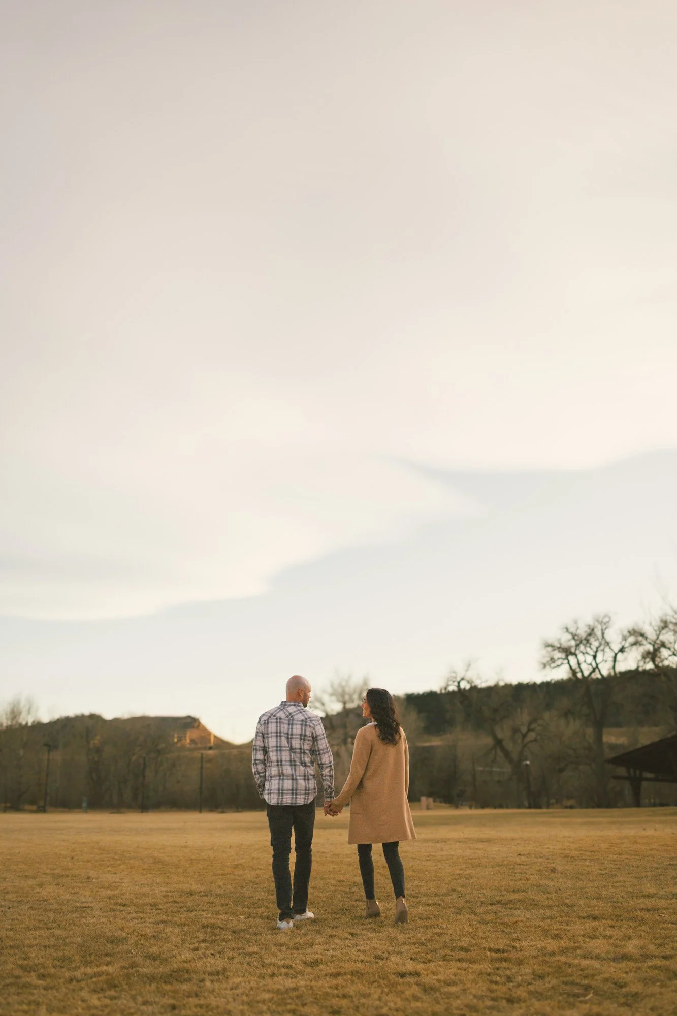 A couple holding hands and walking together on a grassy field during late afternoon or early evening with trees and rolling hills in the background.