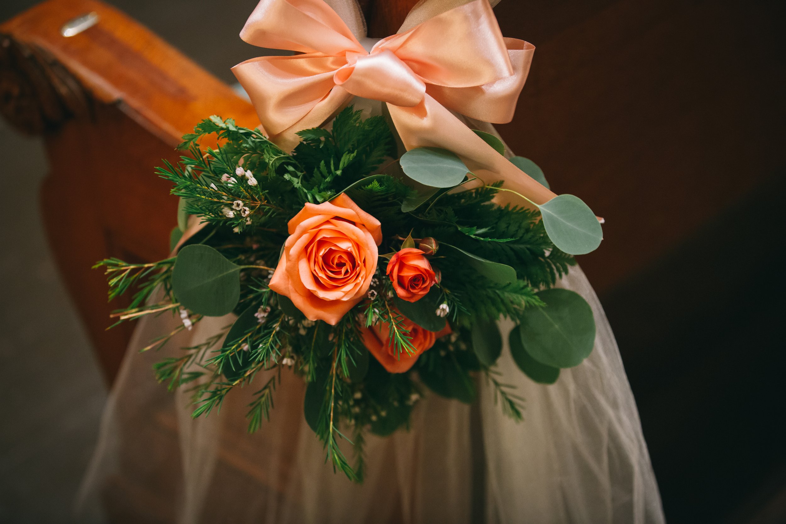 Close-up of a bouquet containing orange roses, greenery, and a large pink satin bow, held by a person wearing a wedding dress.