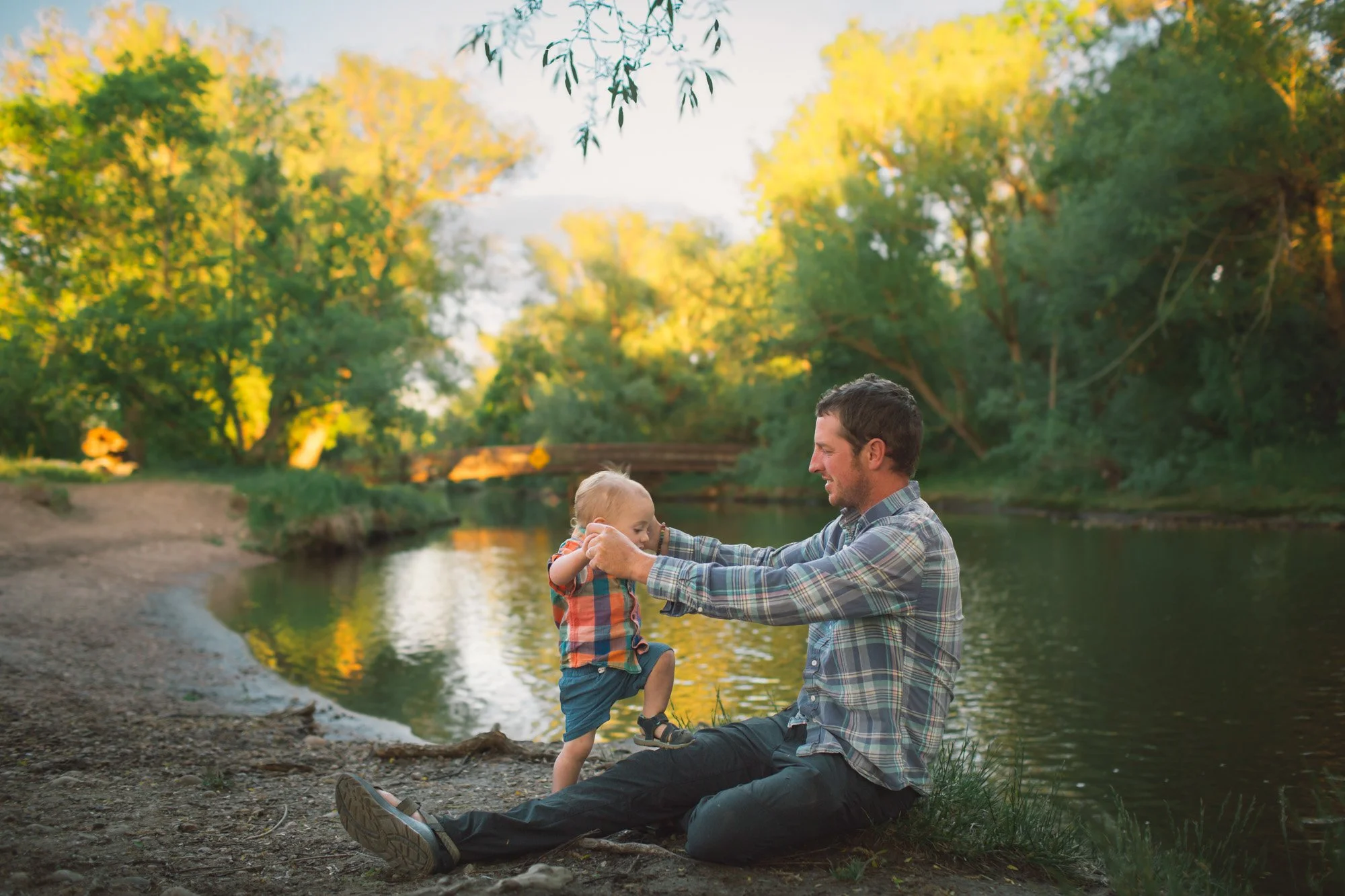 A father and his young son playing by a river in a forest with green and yellow trees during sunset.