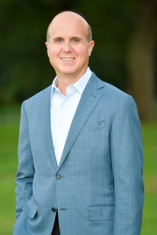 A smiling man in a light blue suit jacket and white dress shirt, standing outdoors with blurred greenery in the background.