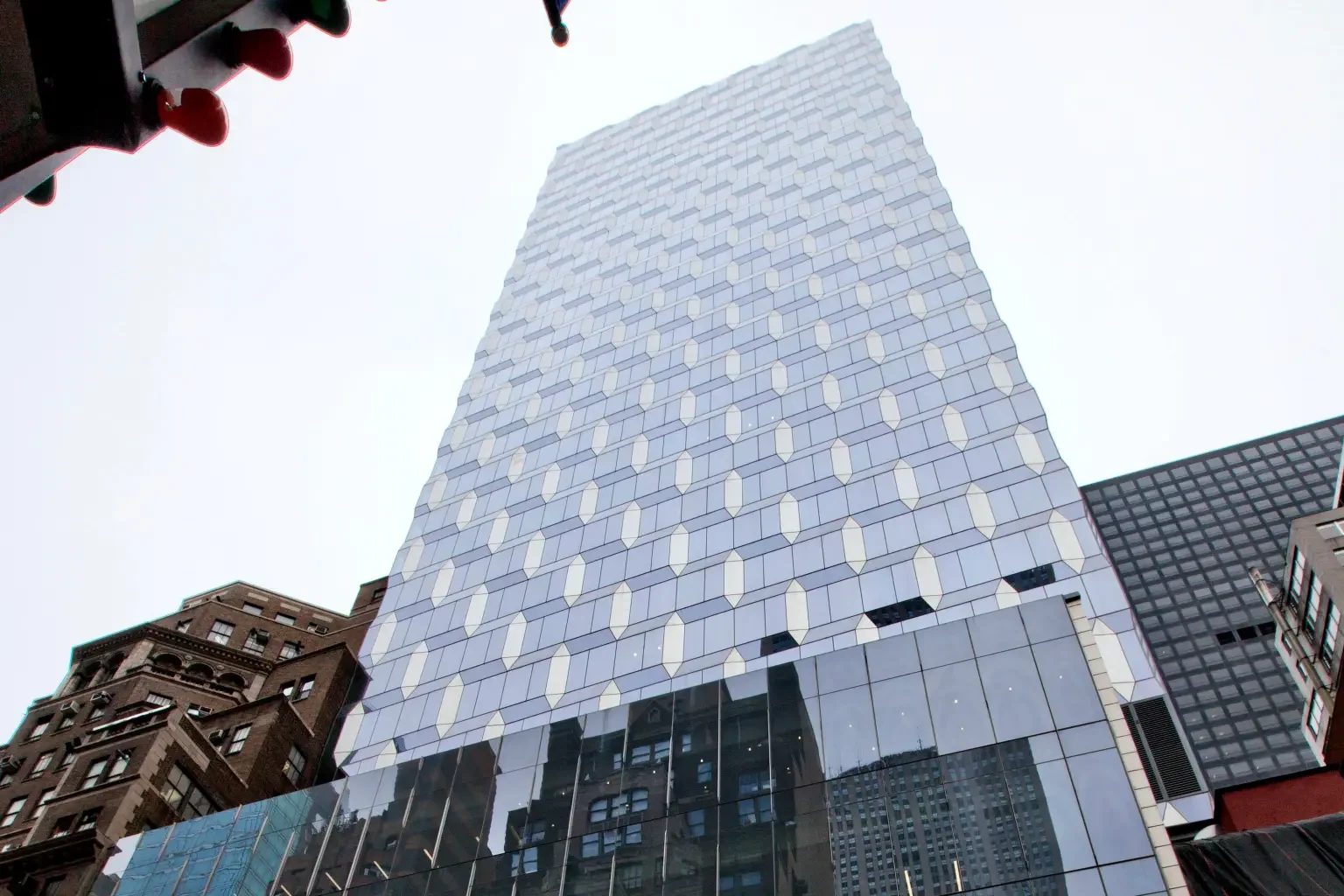 Tall modern glass skyscraper seen from below with a pattern of white accents on its facade, surrounded by other buildings in an urban setting.