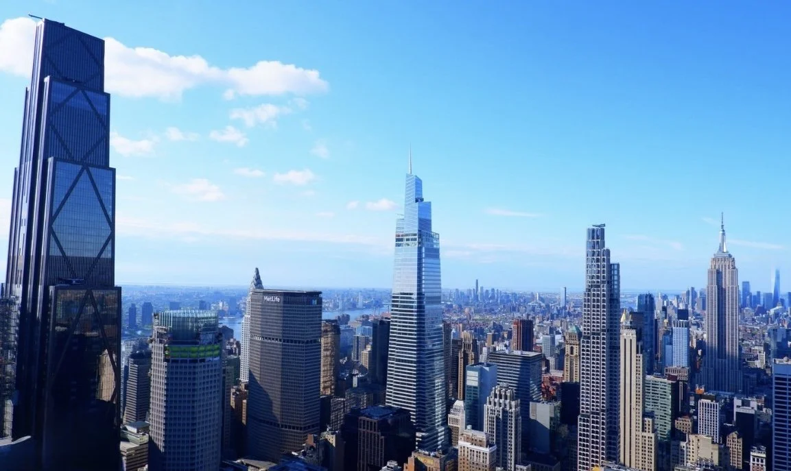 A cityscape of New York City featuring skyscrapers and the Empire State Building in the distance on a bright, sunny day.