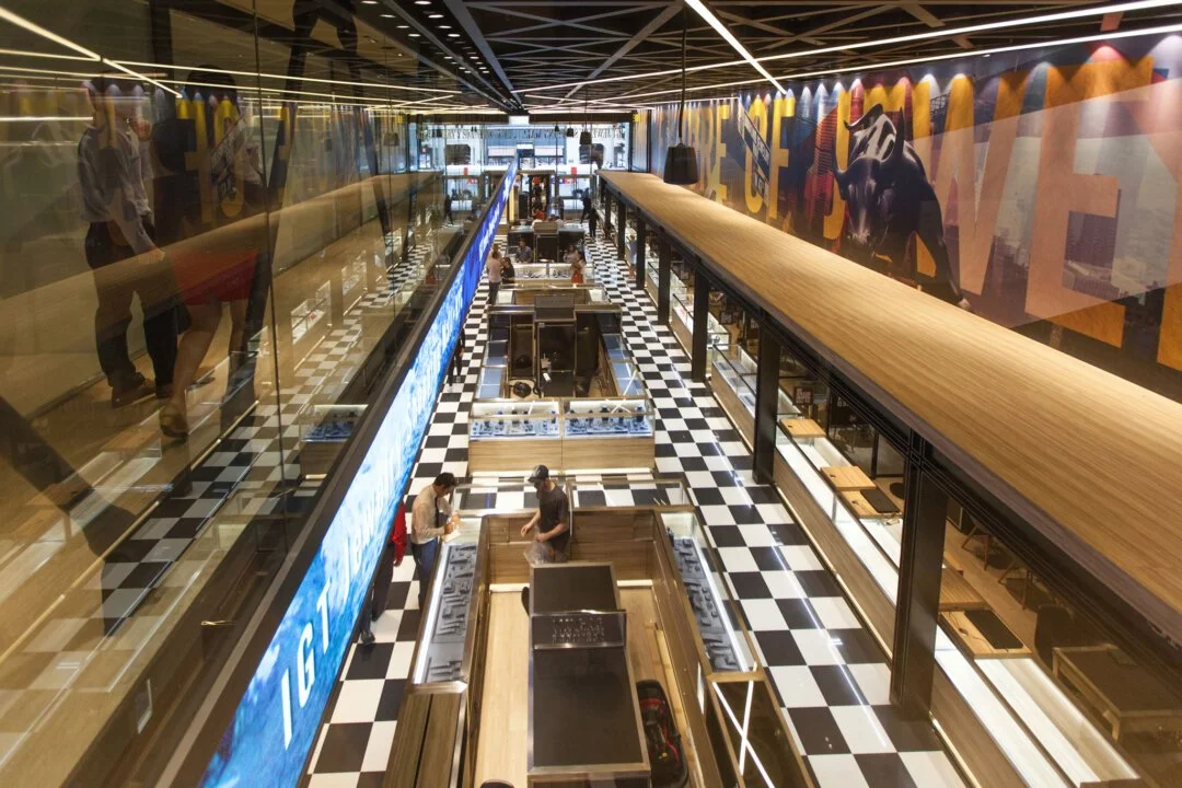 Interior of a modern shopping mall with a black-and-white checkered floor, wooden display counters, a large colorful mural on the wall, and an upper level with a wooden railing. There are a few people shopping.