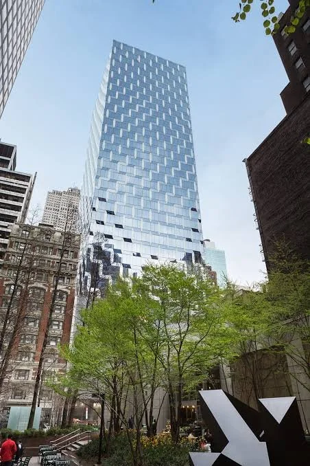 A tall modern glass skyscraper in an urban setting with trees and other buildings around it, viewed from street level.