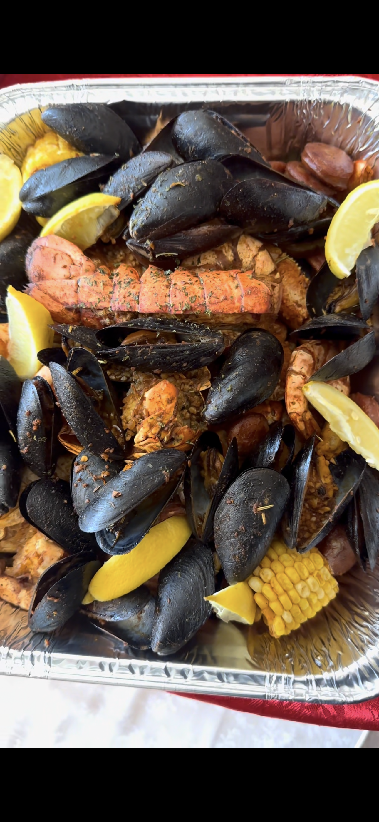 Seafood platter featuring mussels, lobster, shrimp, lemon wedges, corn on the cob, and sausage in a foil tray.