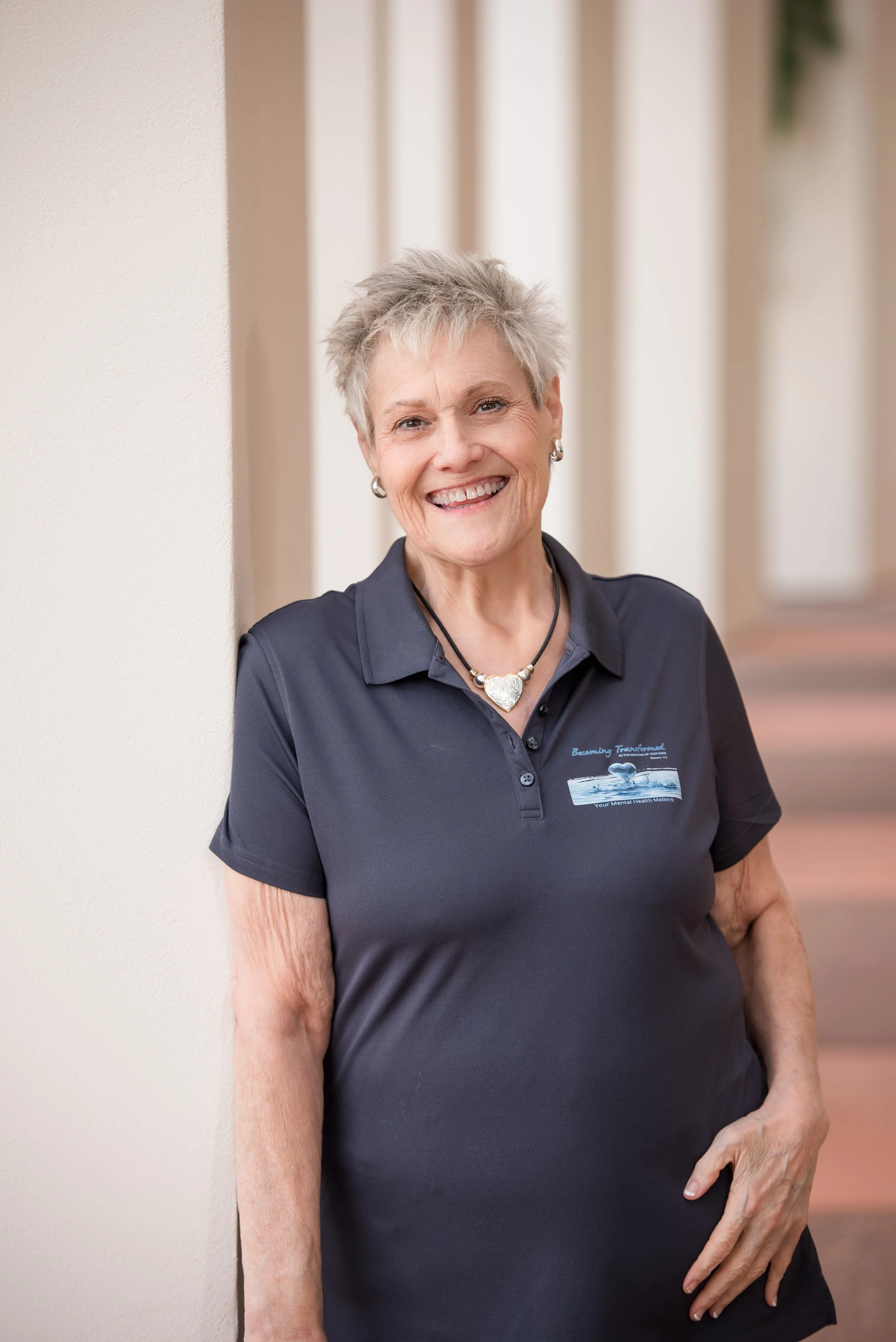 A smiling elderly woman wearing a navy blue polo shirt with a logo that says "Becoming Trustworthy" and a heart-shaped necklace, standing against a beige wall with vertical wooden blinds in the background.