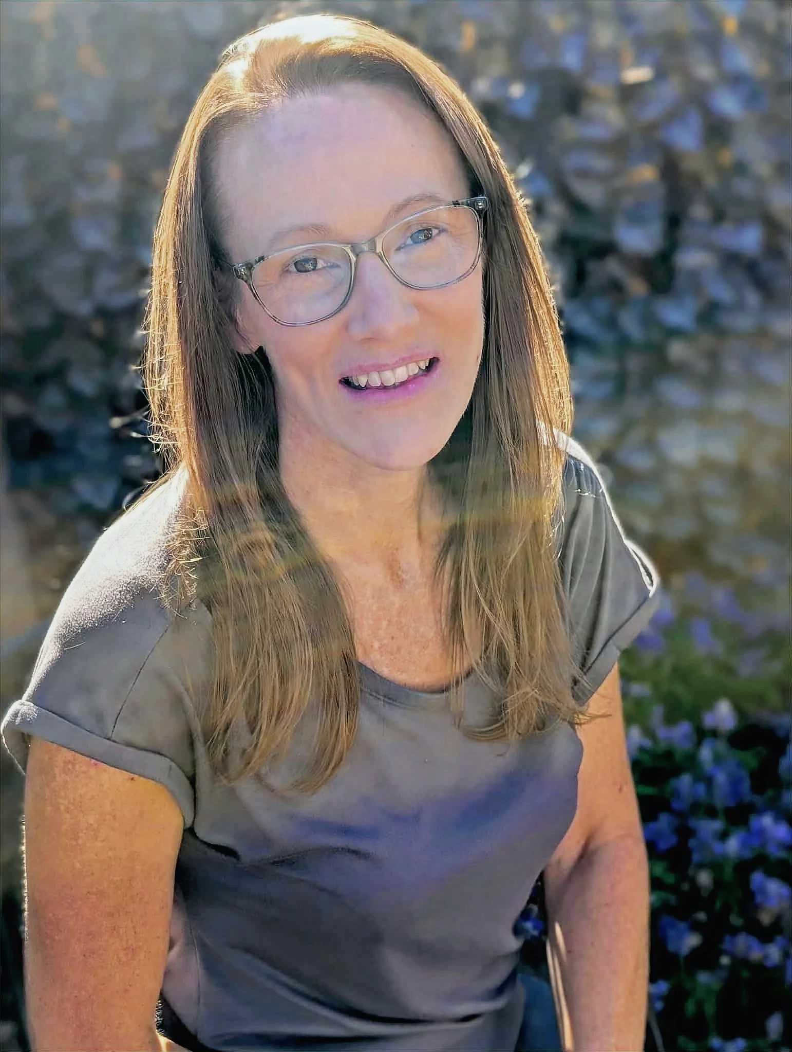 A middle-aged woman with long brown hair, glasses, and a light gray shirt smiling outdoors, with a blurred background of rocks and greenery.