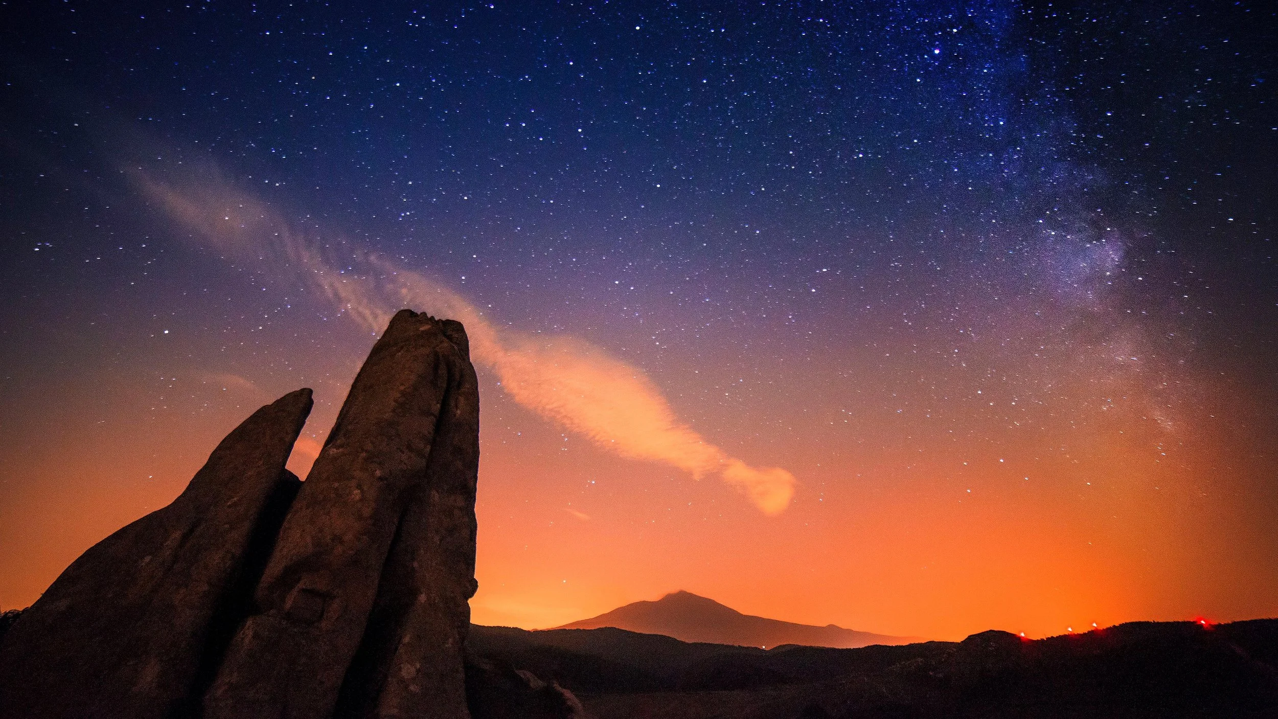 Night sky filled with stars and the Milky Way galaxy, mountain landscape with large rocks in the foreground, and a reddish-orange glow near the horizon.