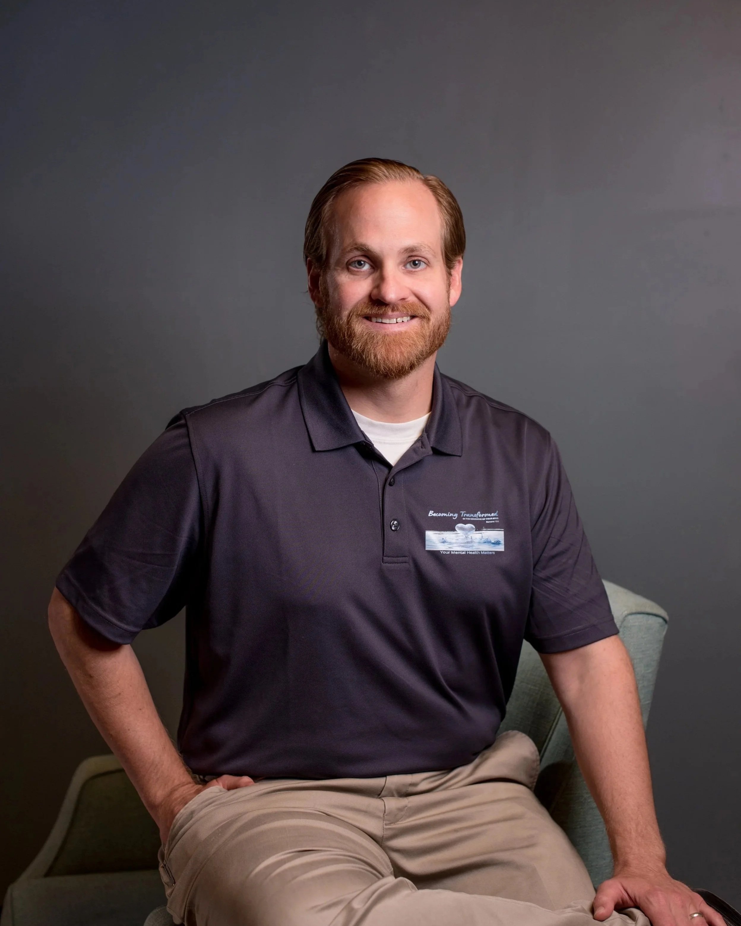 A man with light skin, reddish-brown hair, and a beard, smiling, wearing a dark polo shirt with a badge and beige pants, seated on a chair with a gray background.