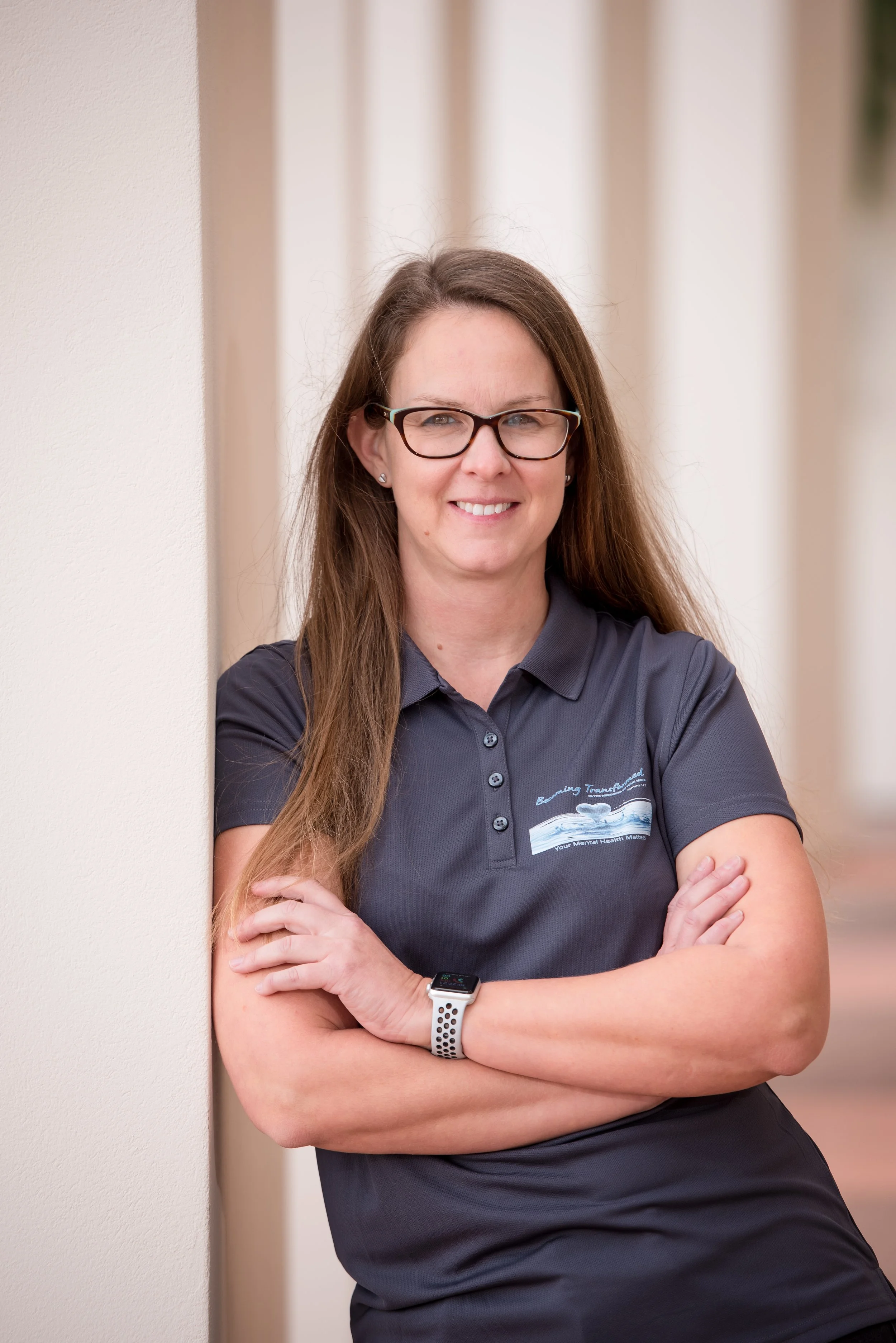 A woman with long brown hair and glasses, smiling with arms crossed, standing against a beige wall and curtains. She is wearing a dark gray collared shirt with a logo.