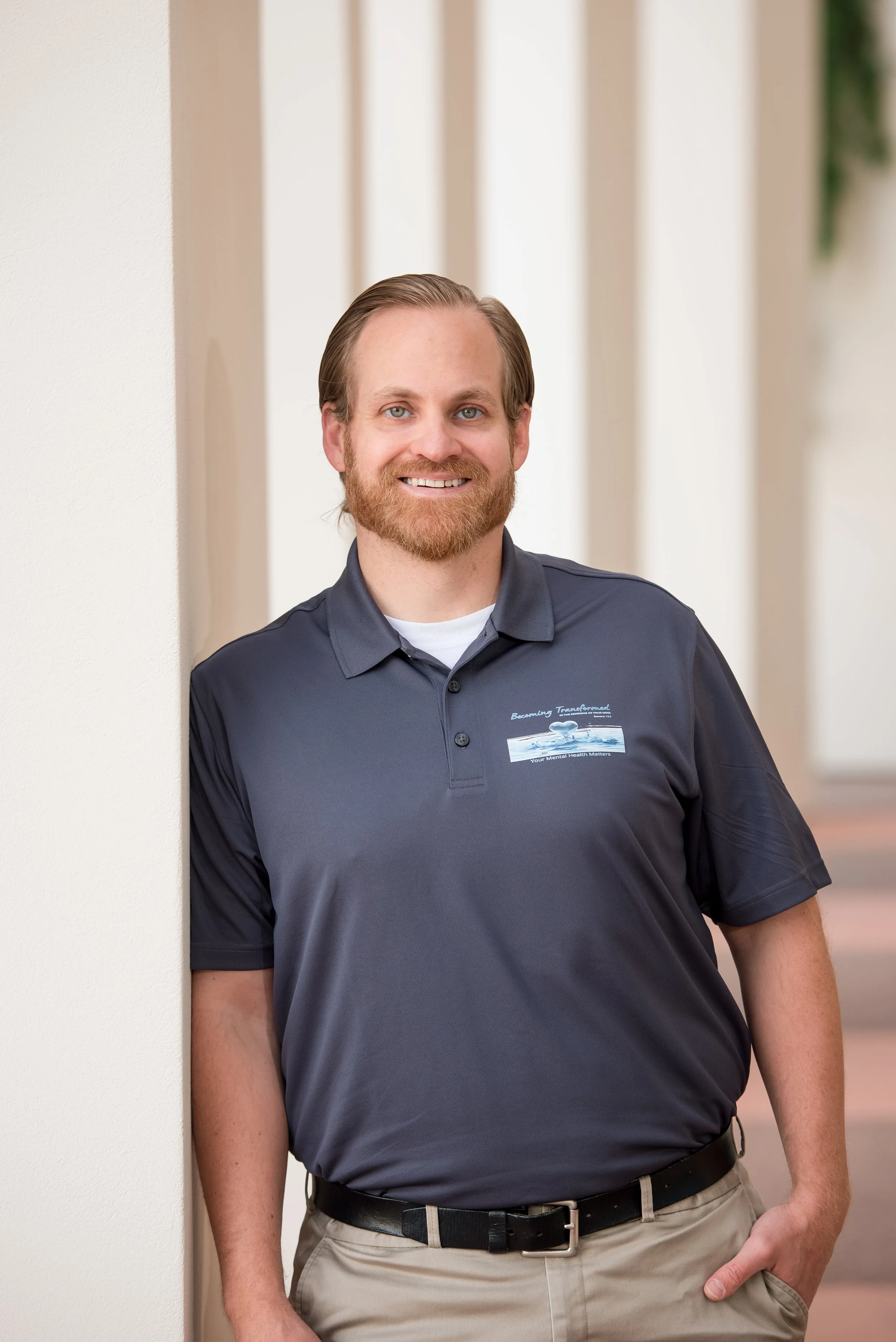A man with a beard and blonde hair smiling, wearing a navy blue polo shirt with a logo, standing against a wall with a blurred background.