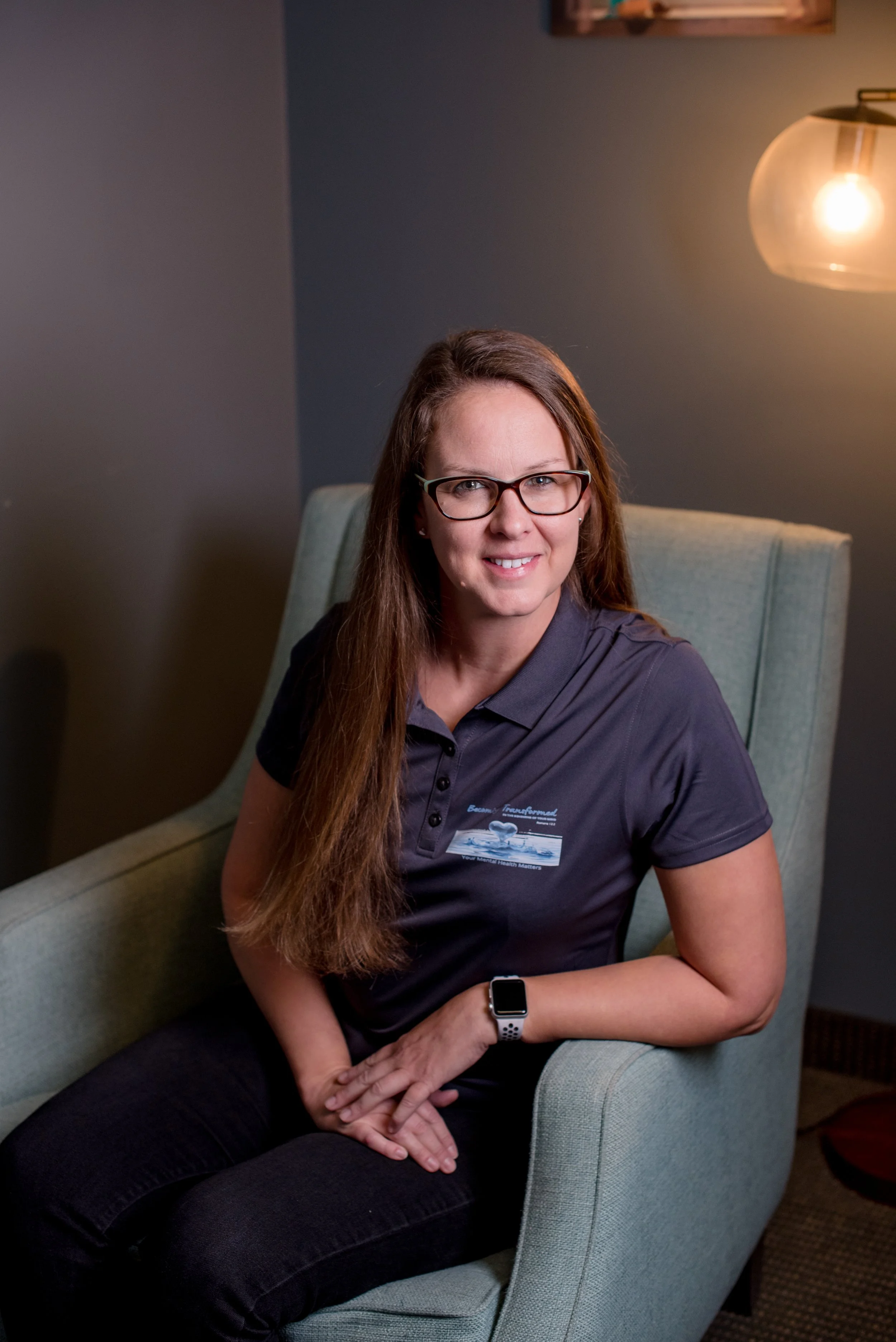 A woman with long brown hair and glasses sitting in an armchair, wearing a dark polo shirt with a logo and a smartwatch, in a dimly lit room with a wall-mounted light fixture.