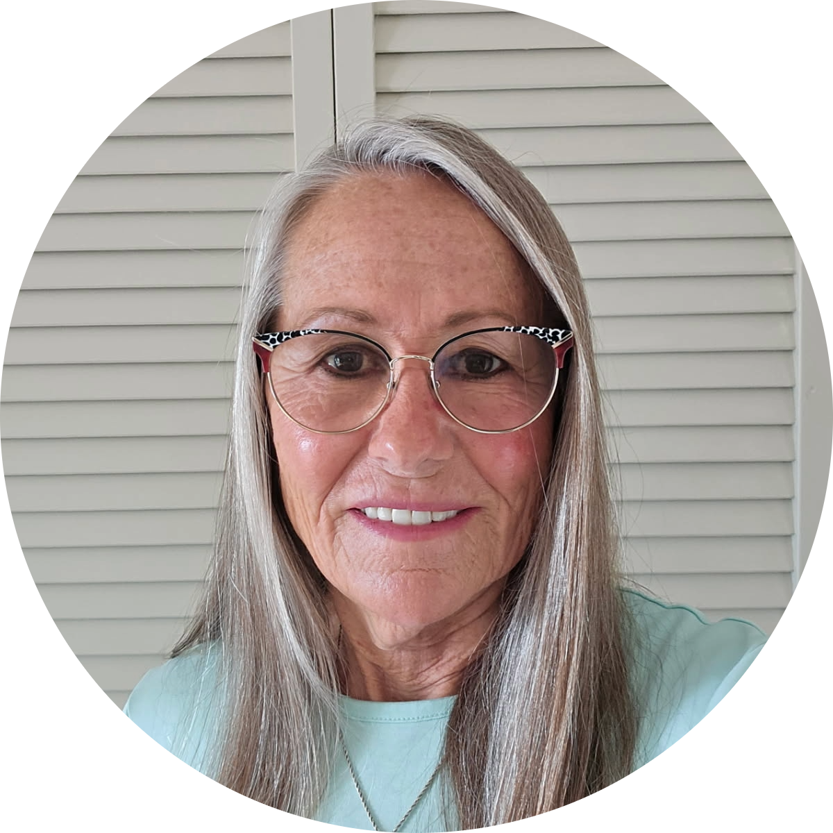 Close-up of smiling older woman with long gray hair, wearing glasses with animal print frames, in front of a beige louvered door.
