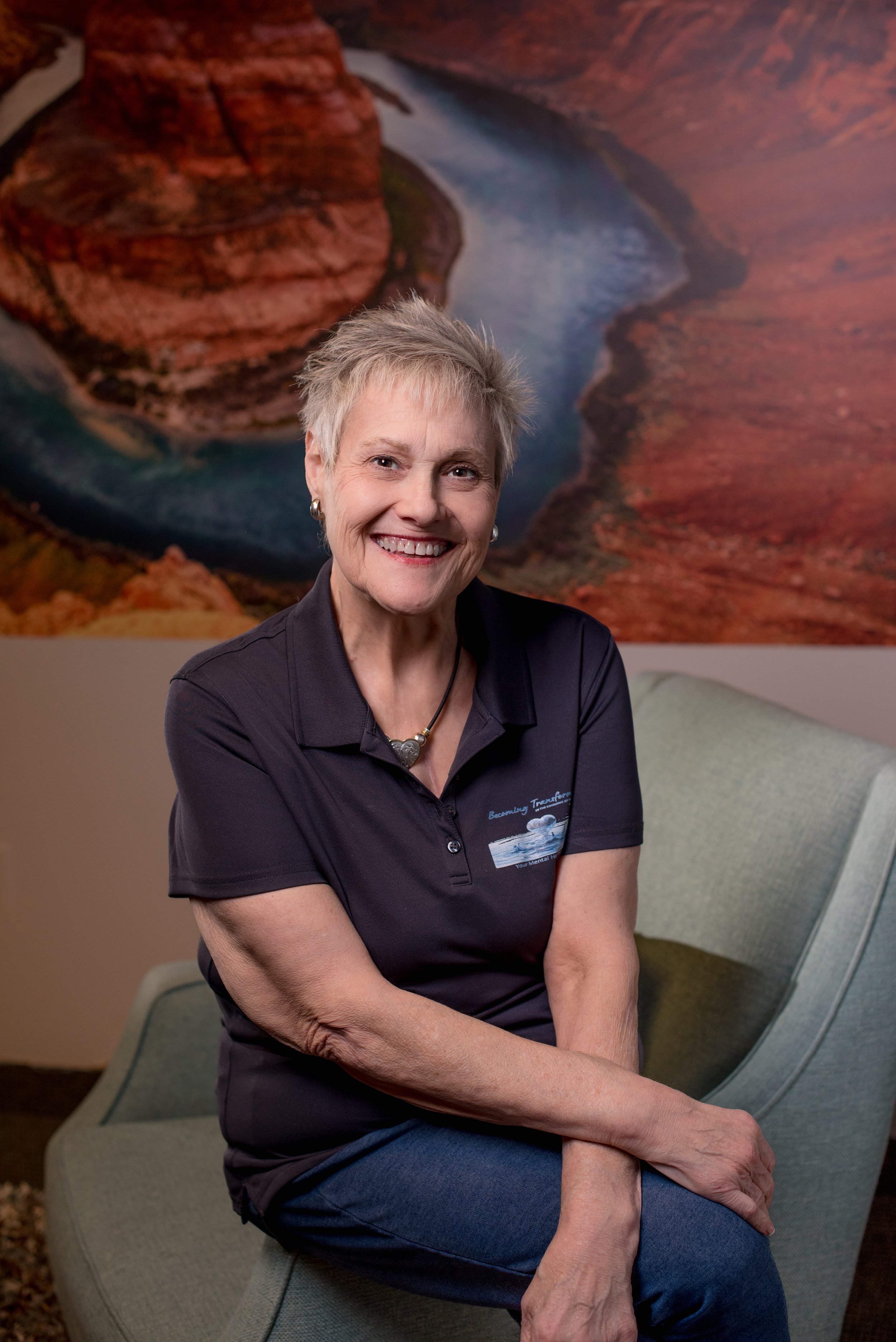 A smiling older woman with short gray hair sitting on a gray chair with crossed legs, wearing a black polo shirt and blue jeans, in front of a wall with a large landscape painting of a canyon with a river.
