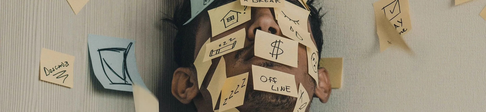 A person's face covered with numerous sticky notes with doodles and words, some of which reveal their eyes, nose, and mouth, as they lay on a wall.