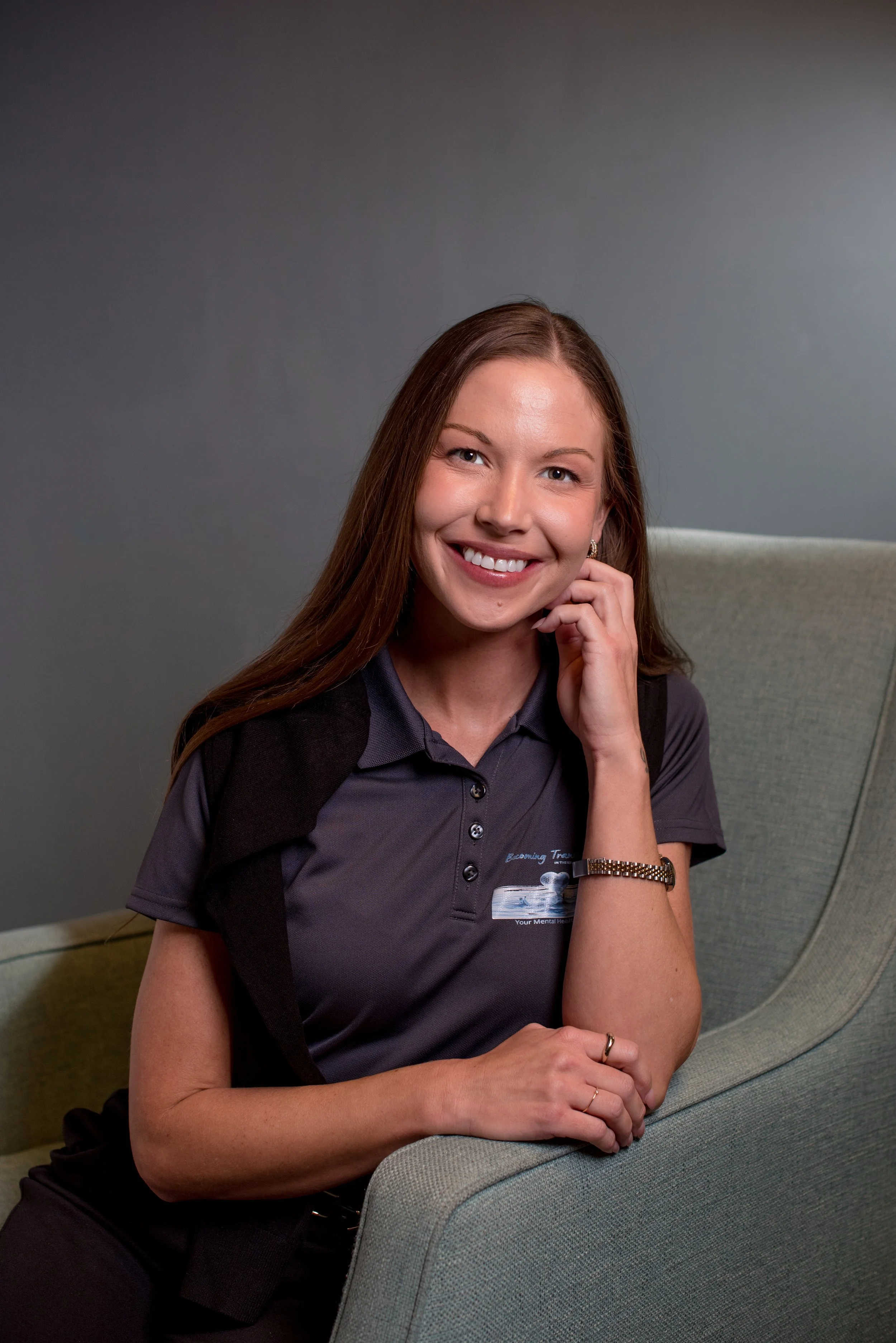 A young woman with long brown hair smiling, wearing a dark gray work shirt with a logo, sitting on a gray and green couch, with her left hand resting on her chin. The background is a plain gray wall.