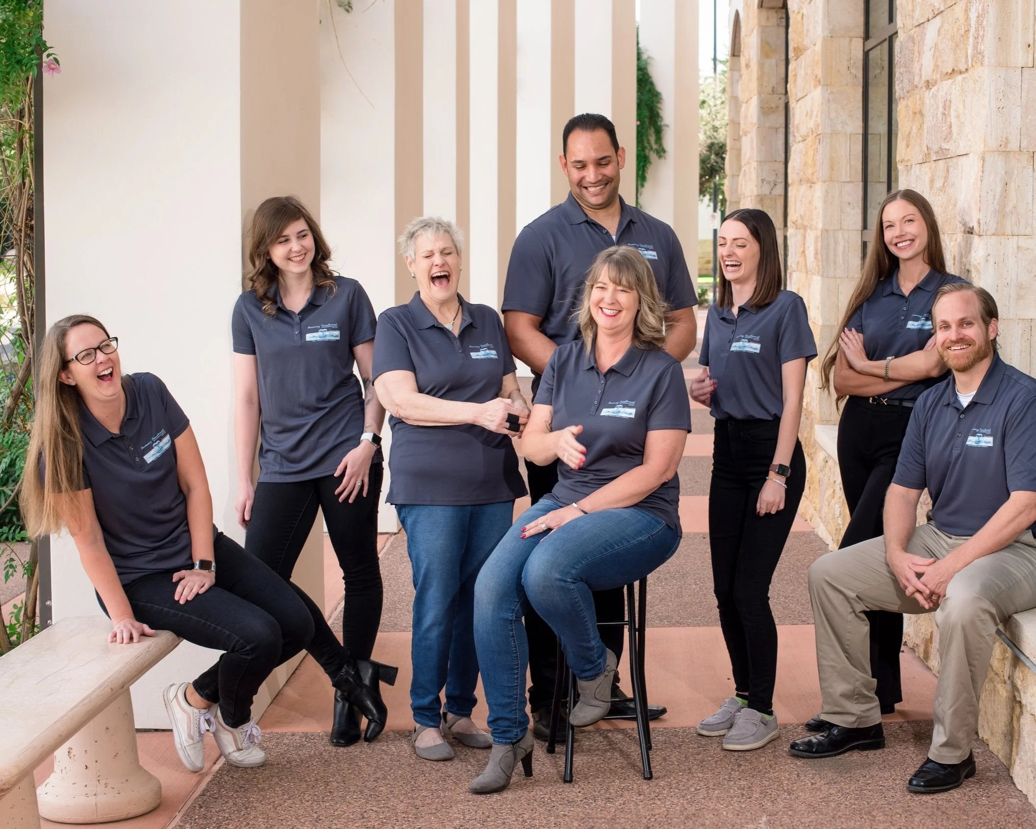 Group of people gathered outdoors, laughing, and smiling. They are wearing matching navy blue polo shirts and are positioned near a stone wall and a bench, with some seated and others standing.