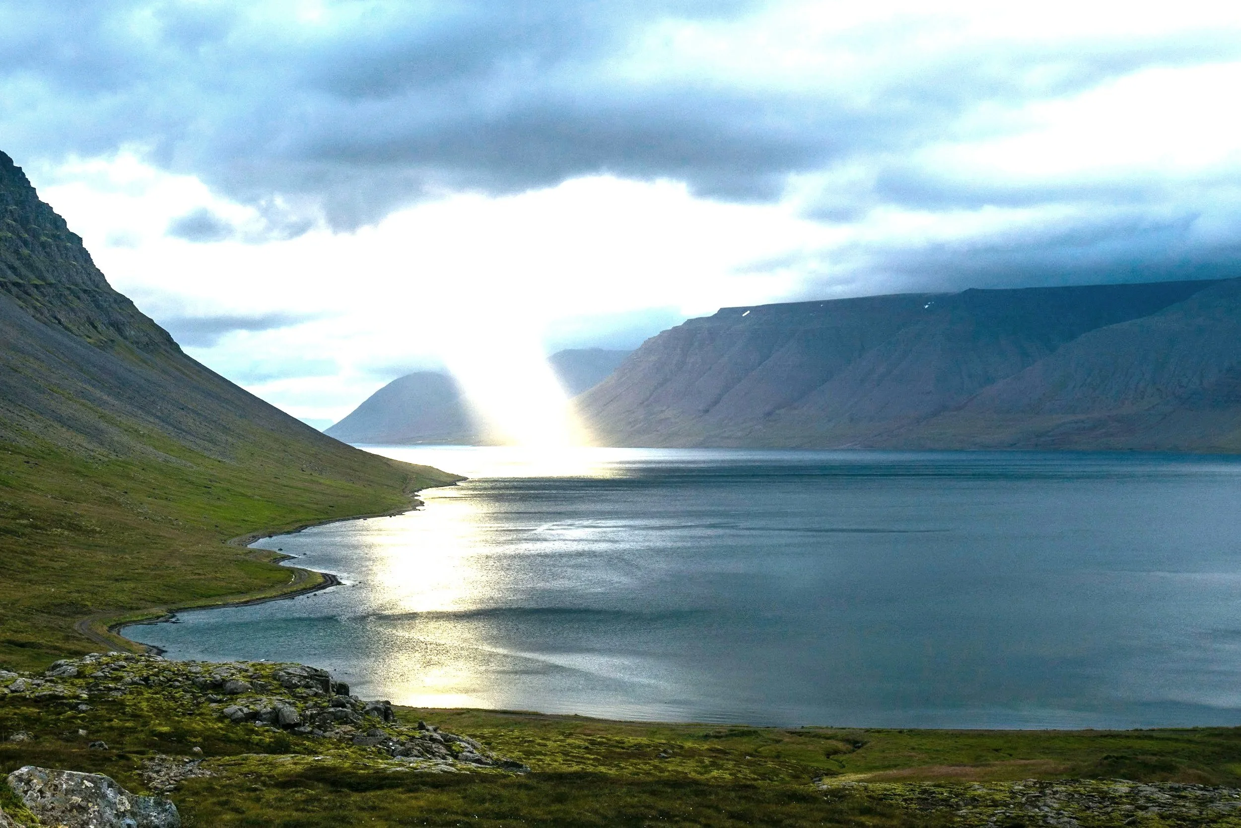 A scenic landscape of a large body of water between green grassy shores and towering mountains under a cloudy sky, with a sunset reflection shimmering on the water.