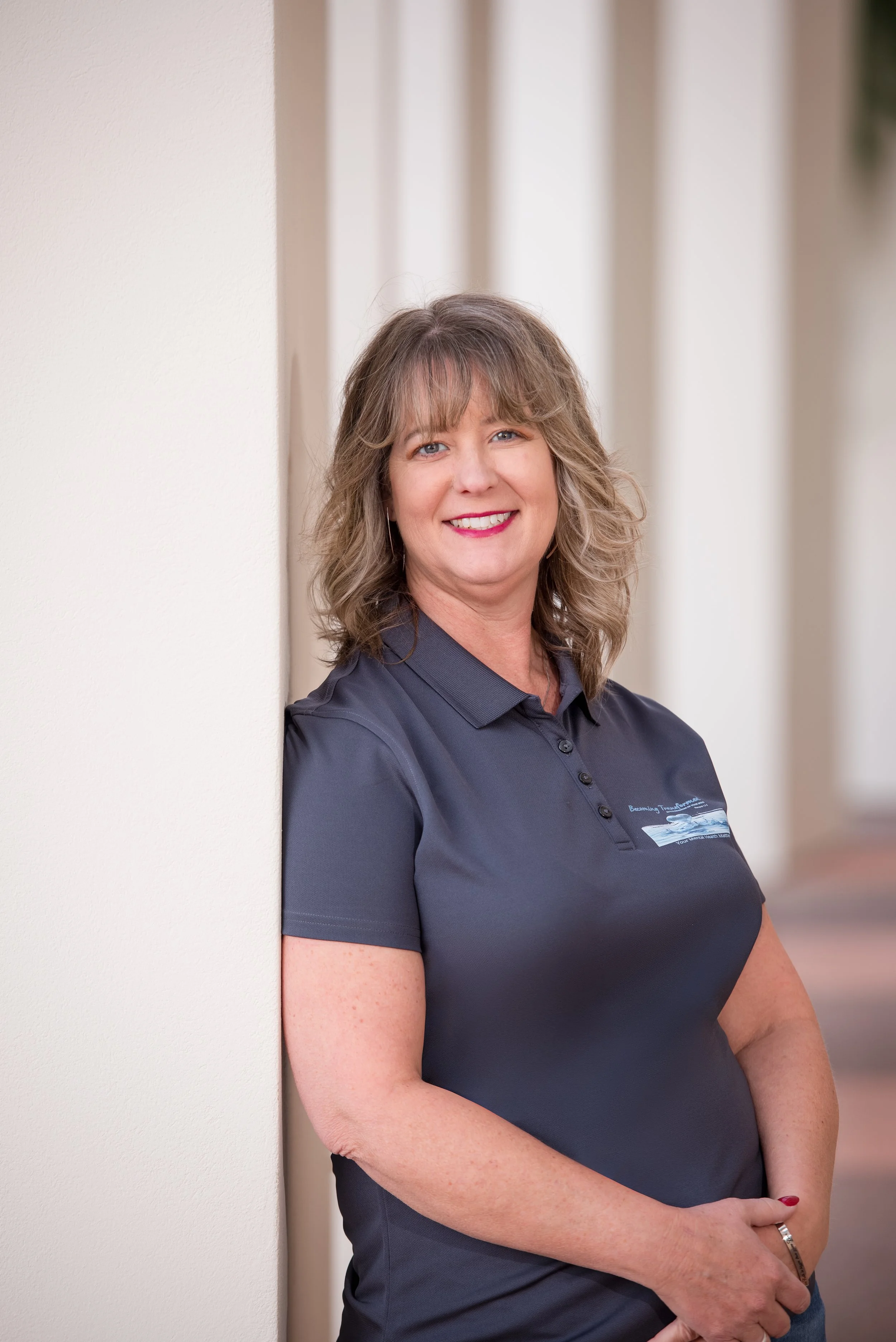 A woman with shoulder-length wavy hair smiling, wearing a dark gray polo shirt, standing next to a white wall.