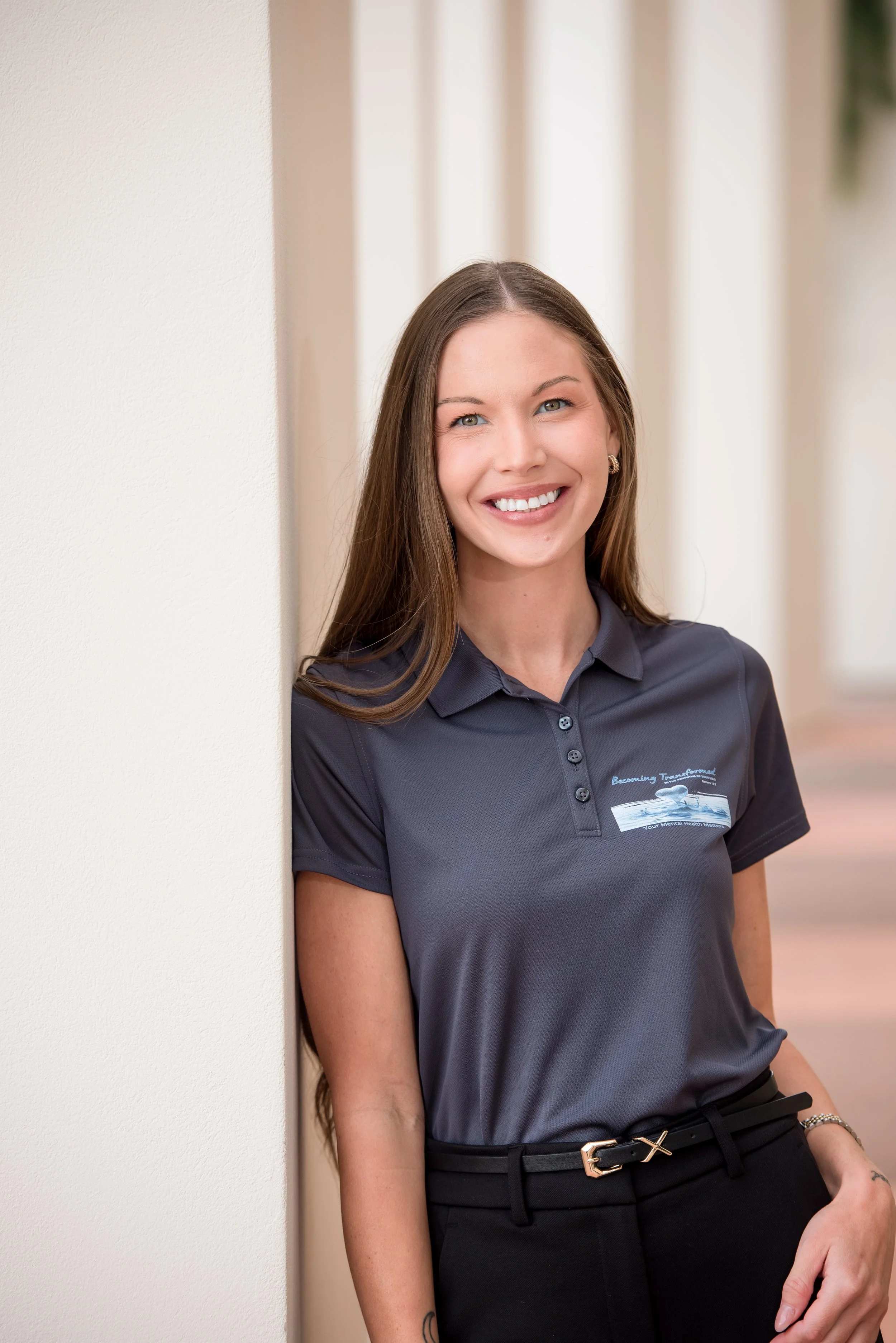 A woman with long brown hair smiling, wearing a dark gray polo shirt with a logo, standing next to a beige wall.