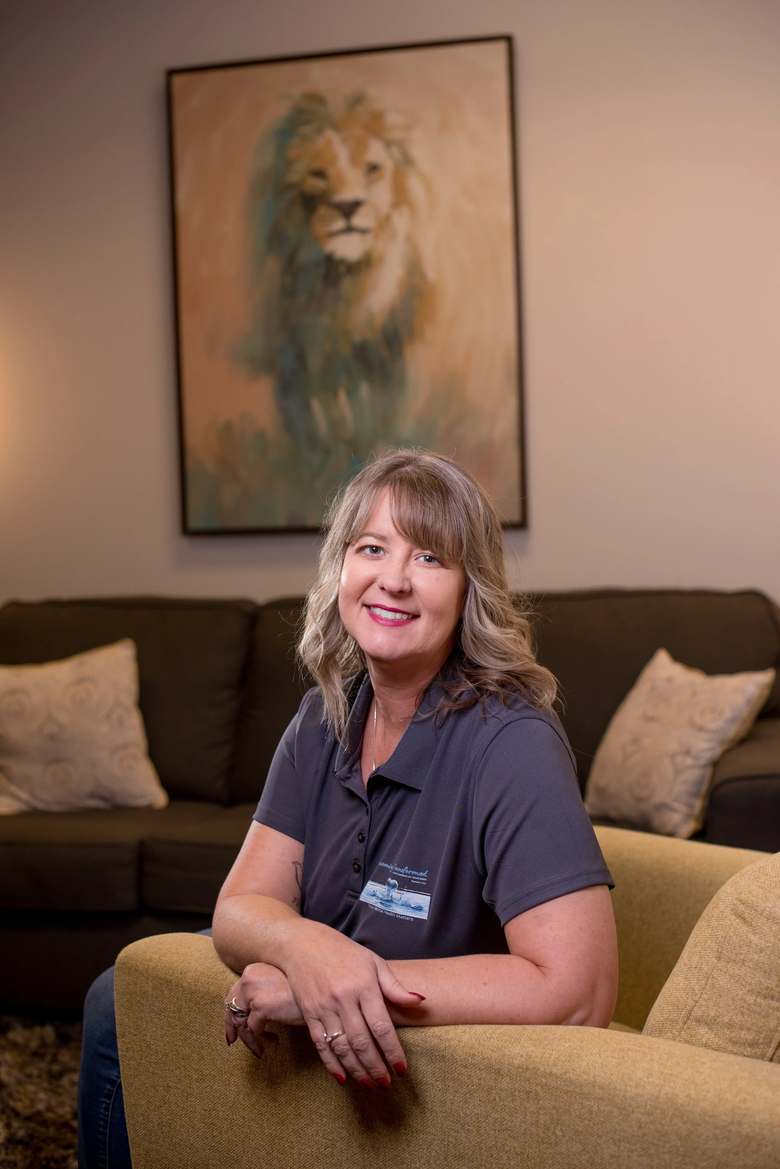 A woman with blonde hair and a dark blue polo shirt sitting on a yellow chair in a living room with a sofa and throw pillows, smiling at the camera. A painting of a lion hangs on the wall behind her.