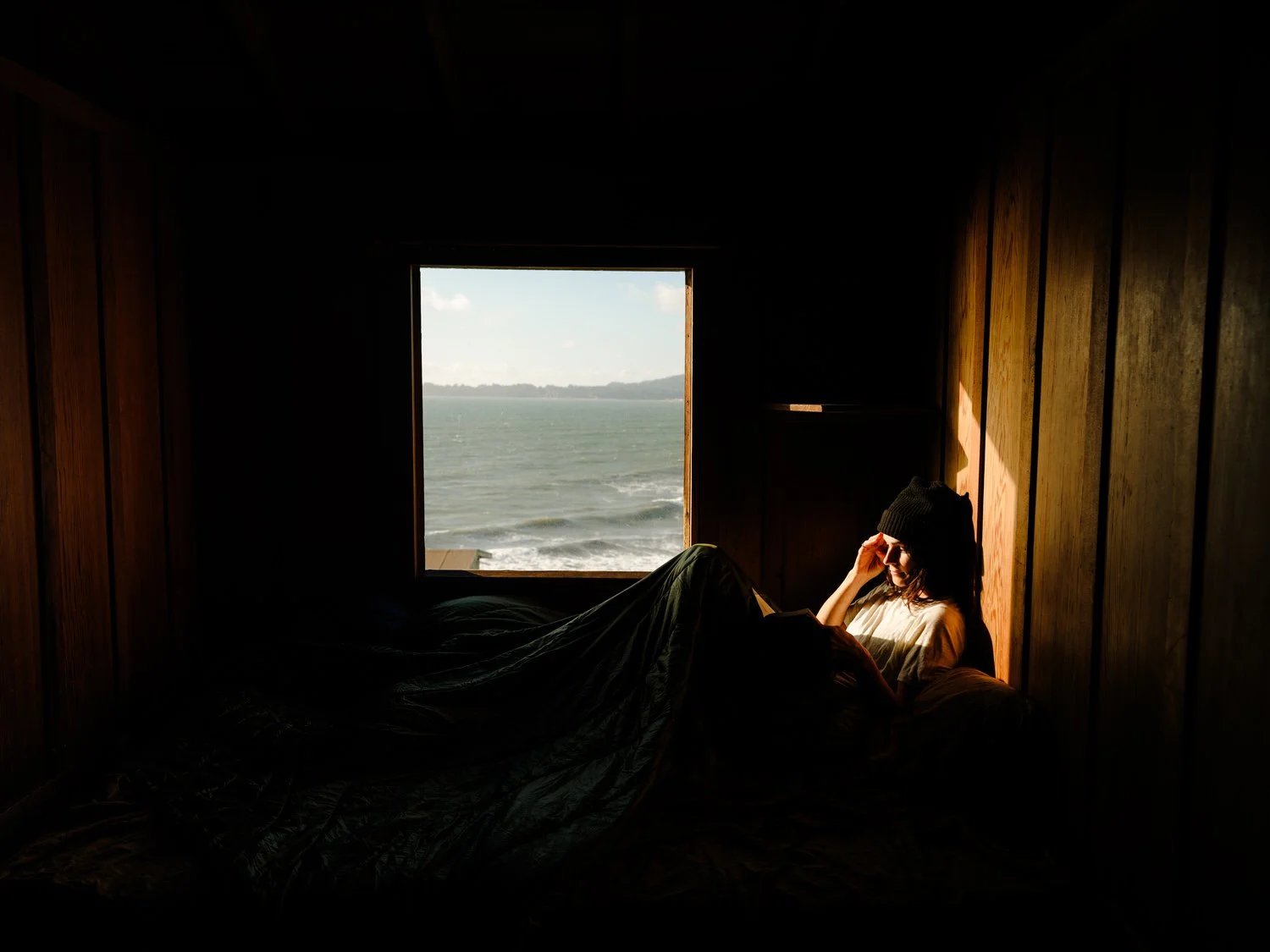 Woman in a beanie hat sitting on a bed inside a wooden cabin, reading a book near a window with ocean view.