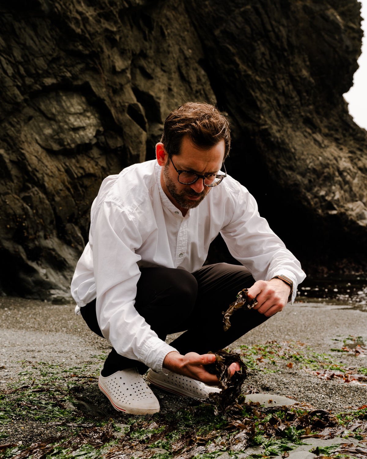 A man in glasses, a white shirt, and white sneakers is crouched on a rocky beach, carefully holding a small crab and examining it. Behind him are large dark rocks and a body of water.