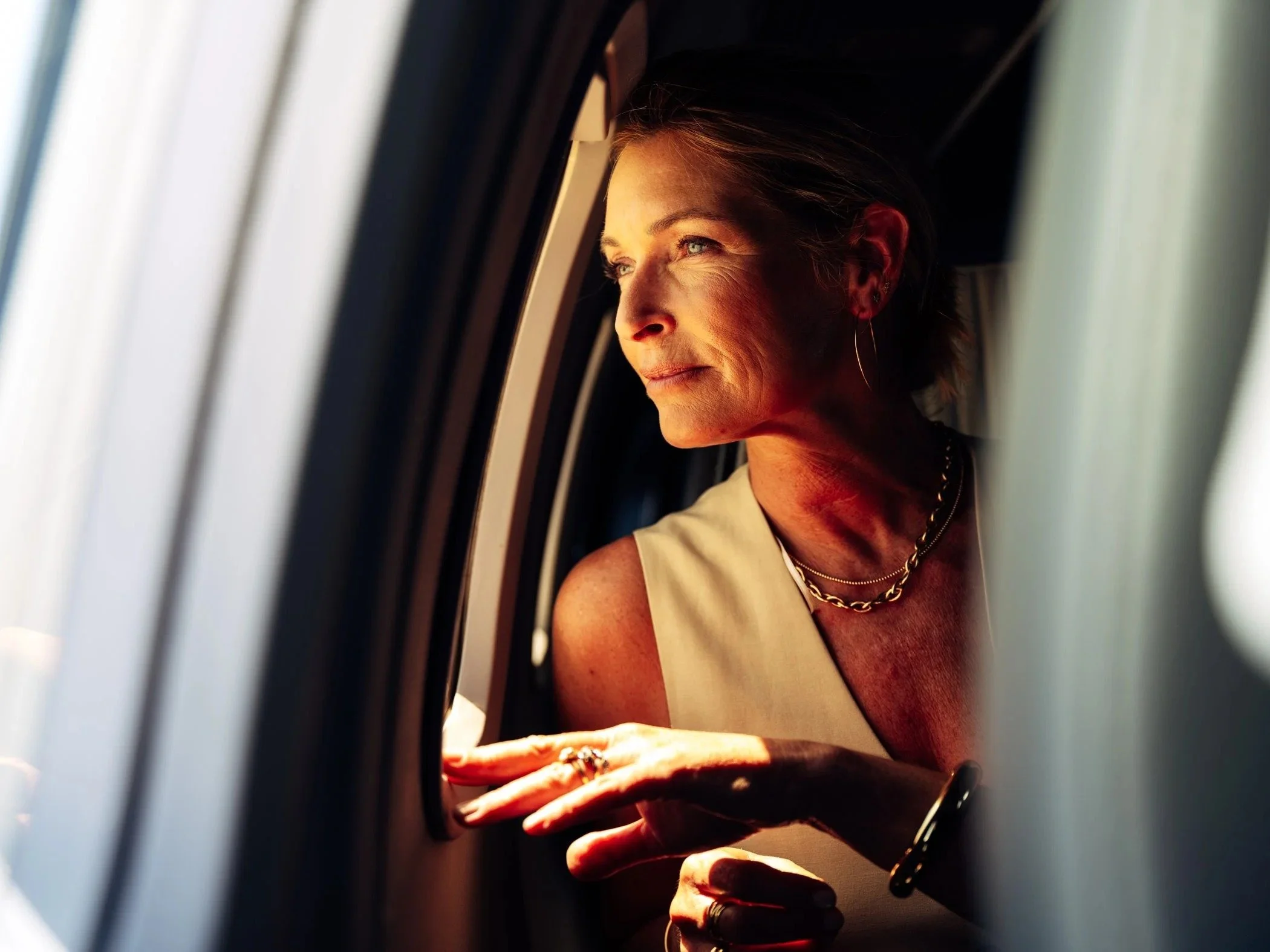 A woman with short hair and blue eyes looking out of a window with soft sunlight illuminating her face, wearing a sleeveless top, a necklace, and earrings.