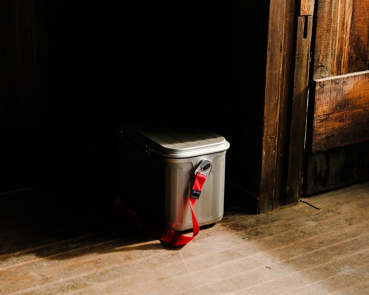 Small container with a red strap attached, placed near a wooden wall in a dimly lit room.