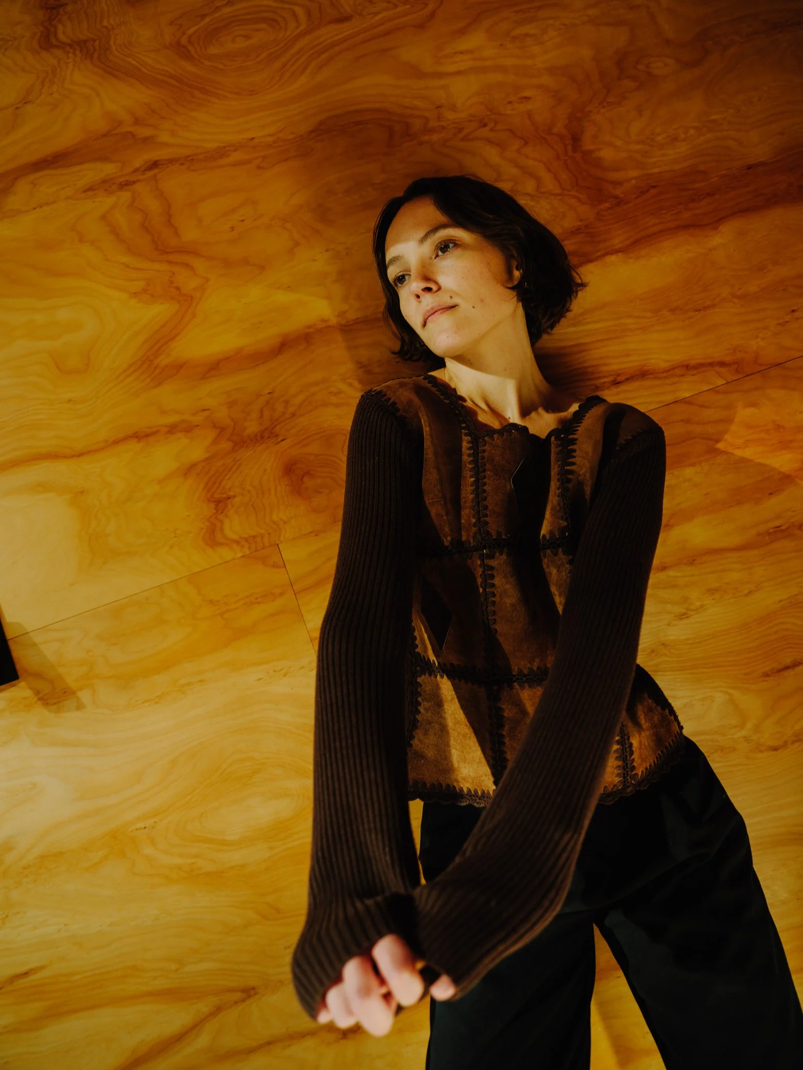 Person with short dark hair, wearing a black and brown patterned jacket, standing in front of a yellowish-brown wood textured background.