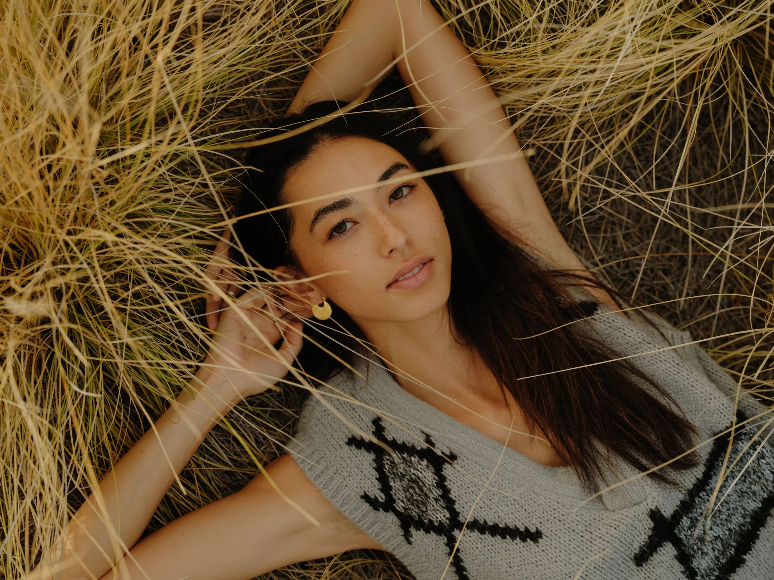 A young woman with dark hair and freckles lying in tall, golden grass, looking at the camera, wearing a gray sweater with black and white pattern and gold crescent moon earrings.