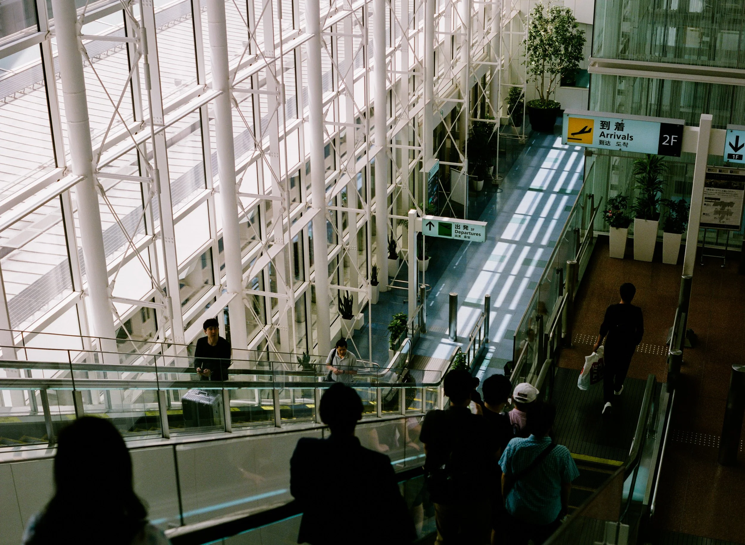 View of an airport interior with large glass windows, showing signs for arrivals and departures, and several travelers walking or standing on escalators.