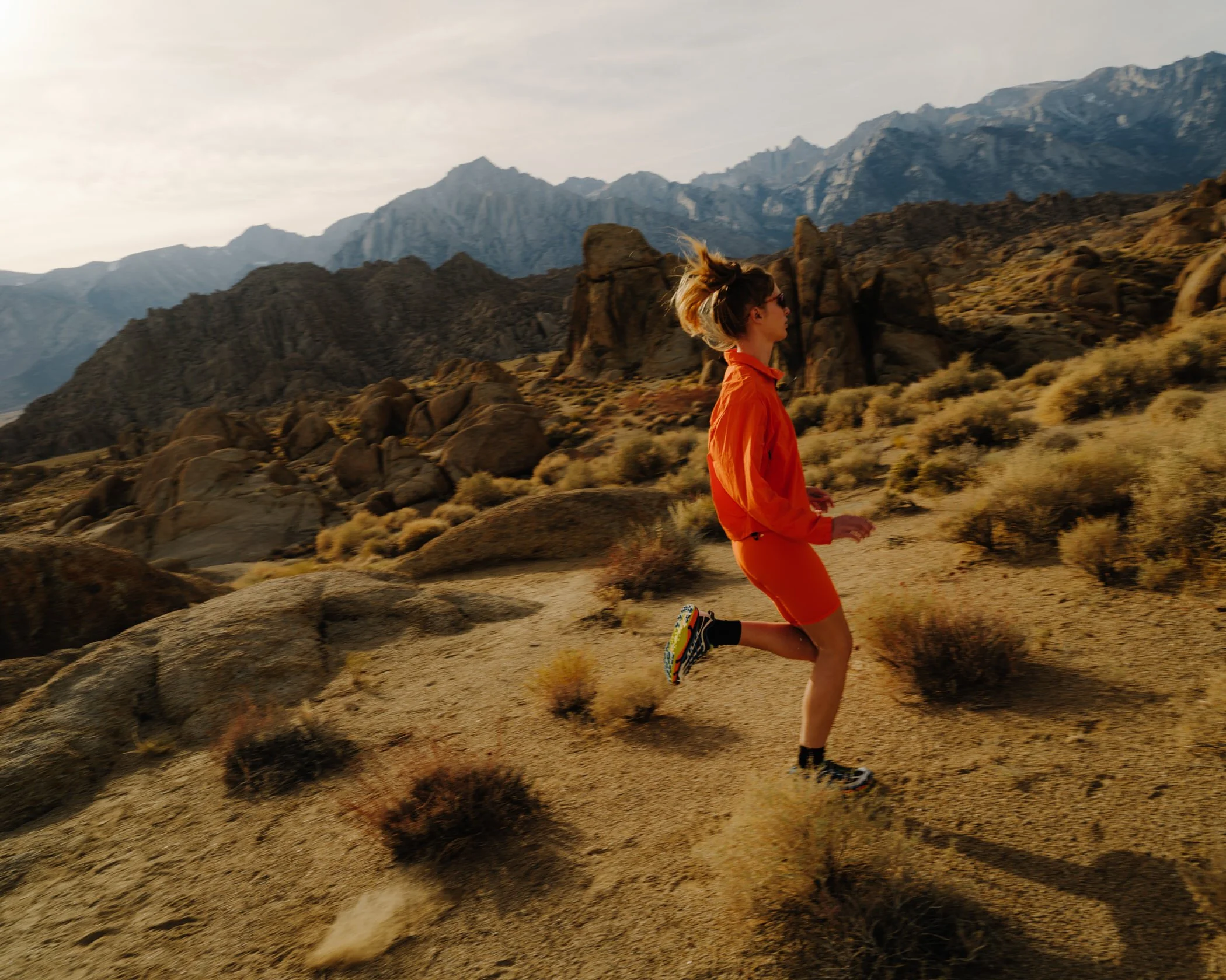 A woman running on a rocky desert trail with mountains in the background, wearing an orange jacket and shorts, and sunglasses.