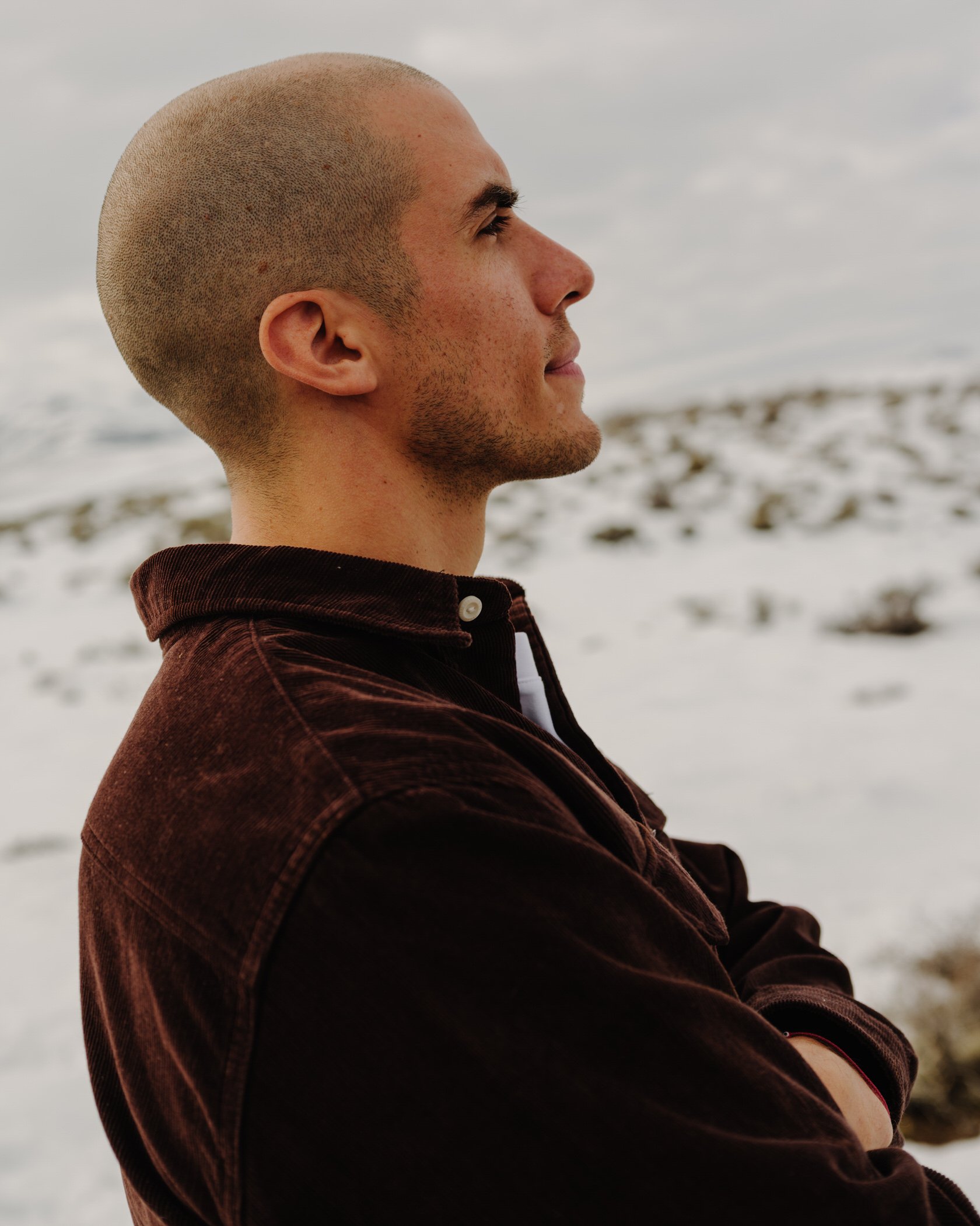 A man with a shaved head and light facial hair relaxing outdoors, possibly on a beach, with blurred sand and waves in the background.
