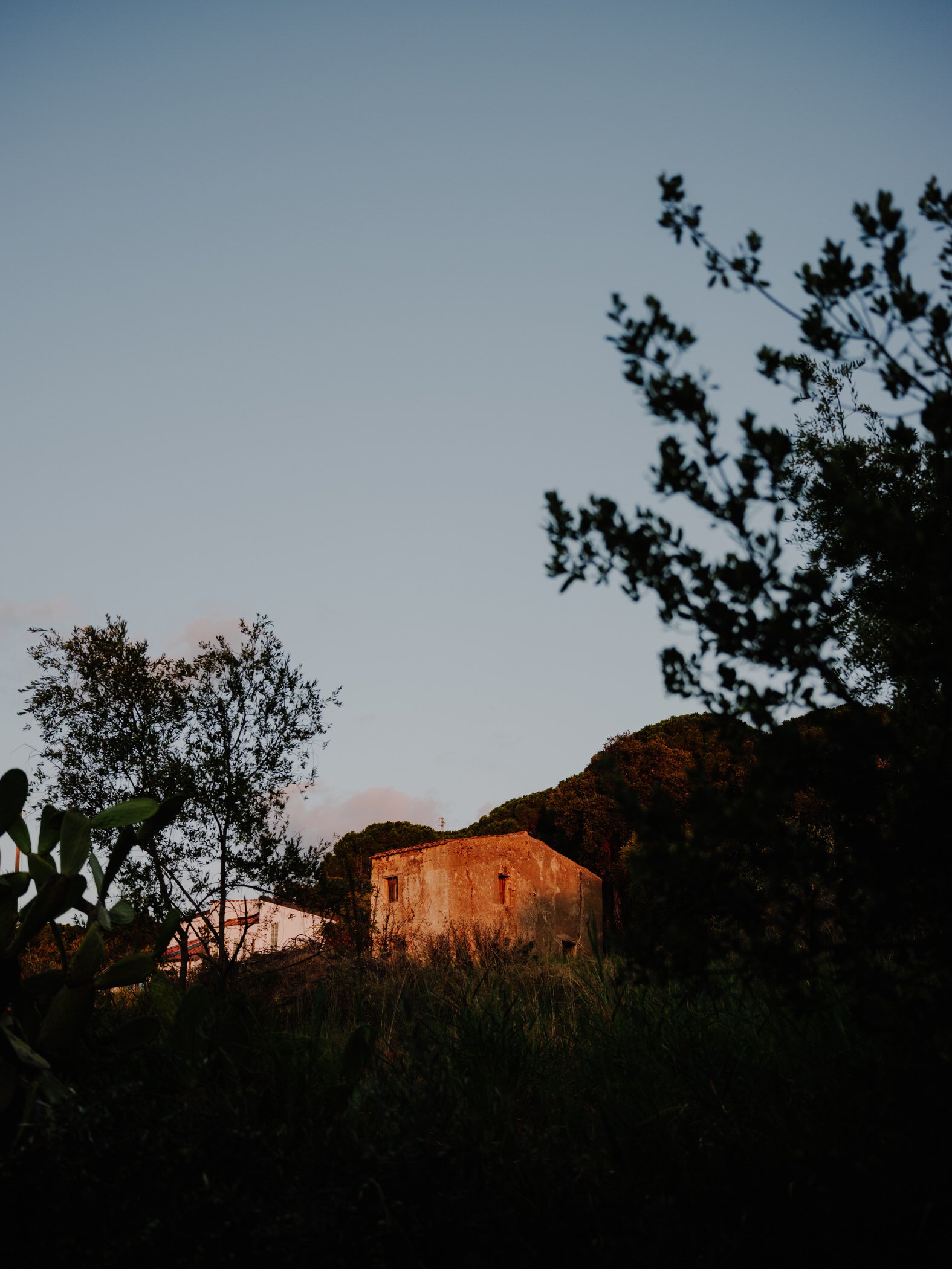 A rural scene at sunset with an old stone house surrounded by trees and bushes, hills in the background, and a clear evening sky.