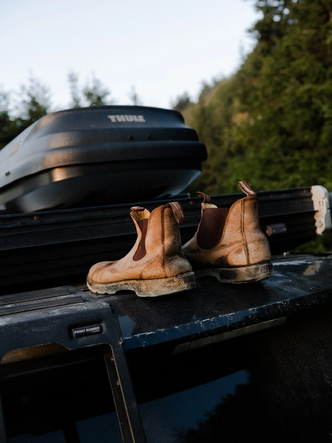 Old muddy leather hiking boots resting on the roof of a vehicle, with a cargo box on top, against a background of trees and sky.