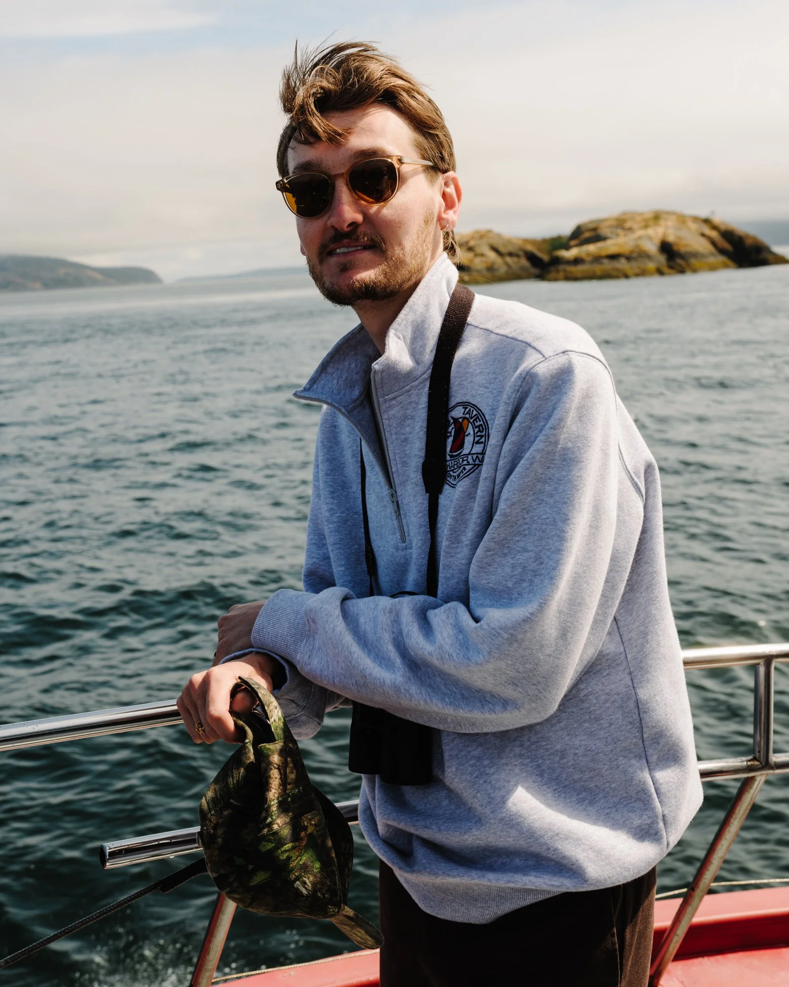 A young man with sunglasses, a beige jacket, and a scruffy beard, holding a fishing line with a fish, standing on a boat with water and islands in the background.