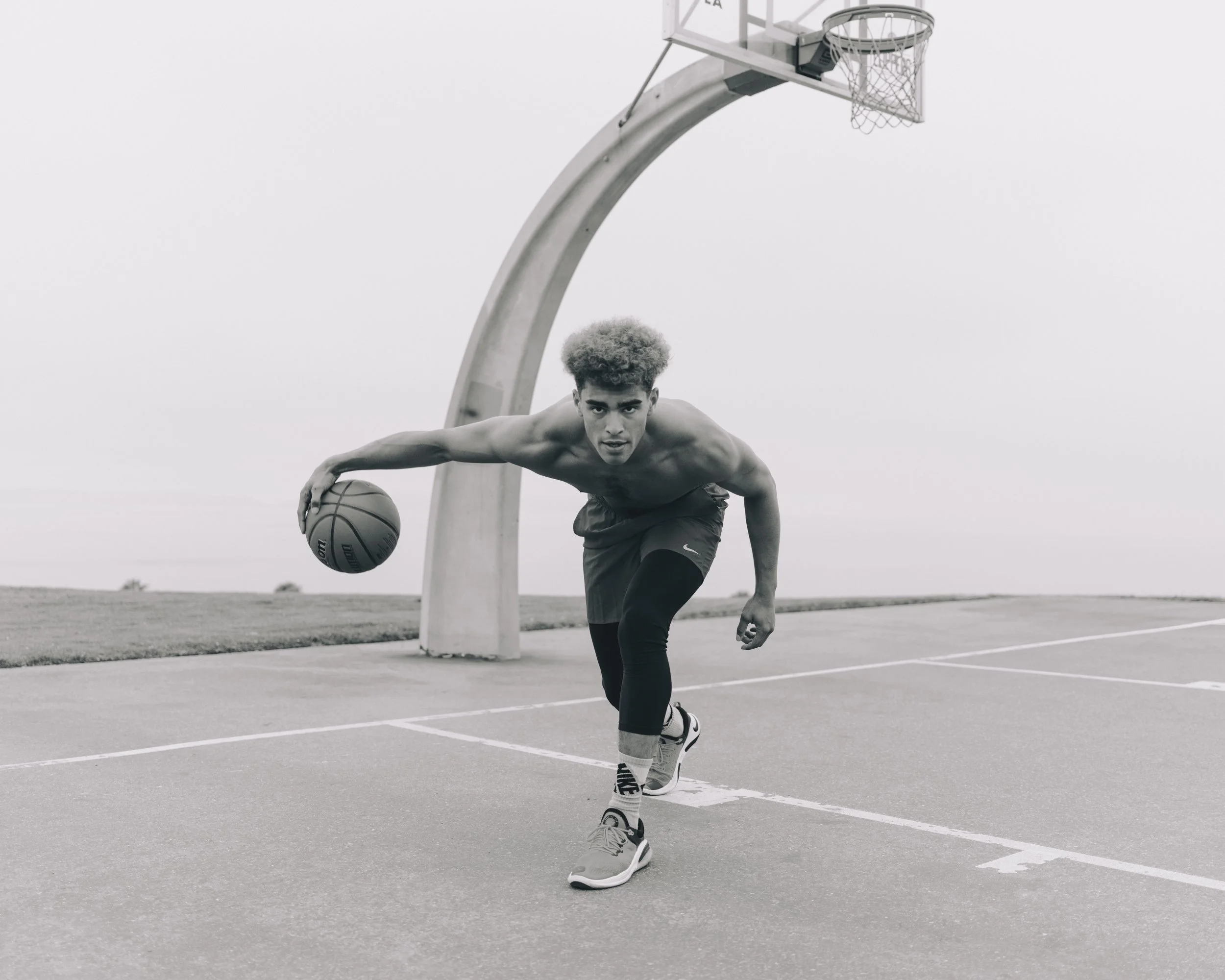 A young man with curly hair and athletic build playing basketball outside on a court. He is shirtless, wearing black athletic pants, Nike sneakers, and striped socks, dribbling a basketball, with a basketball hoop in the background.