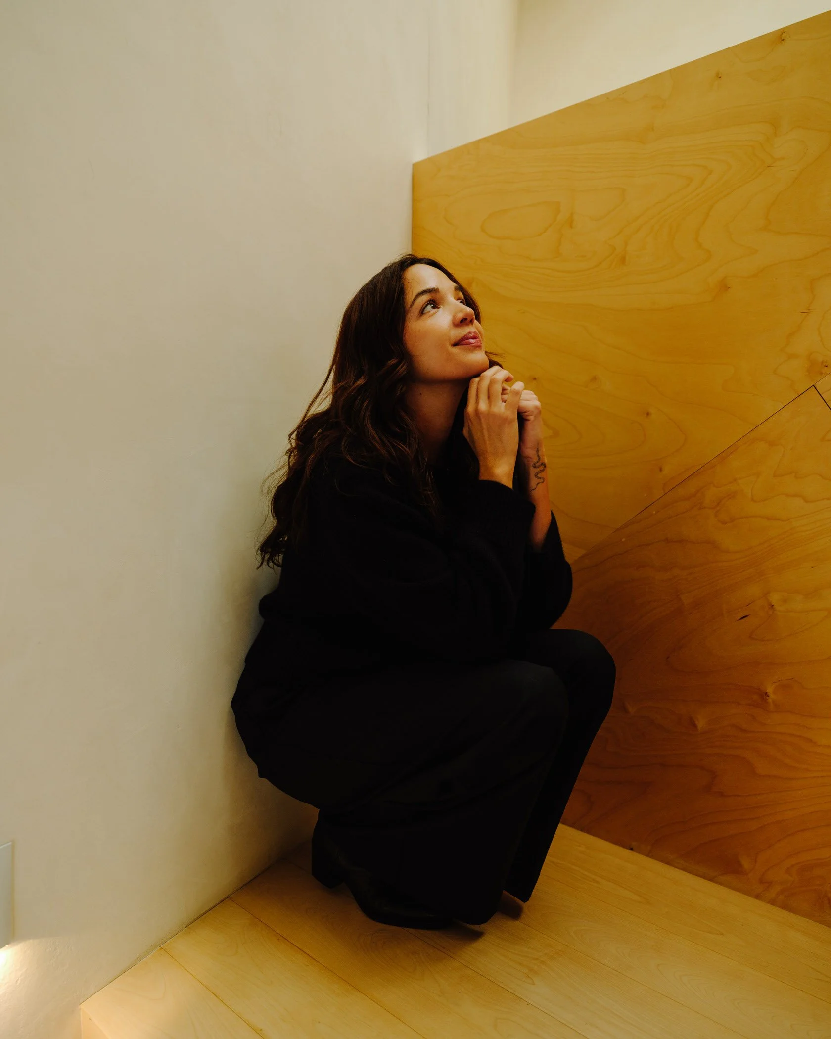 A woman with long, wavy dark hair, sitting on the floor with her hands clasped near her chin, looking upward and smiling, in a minimalistic room with a light-colored wall and wooden furniture.