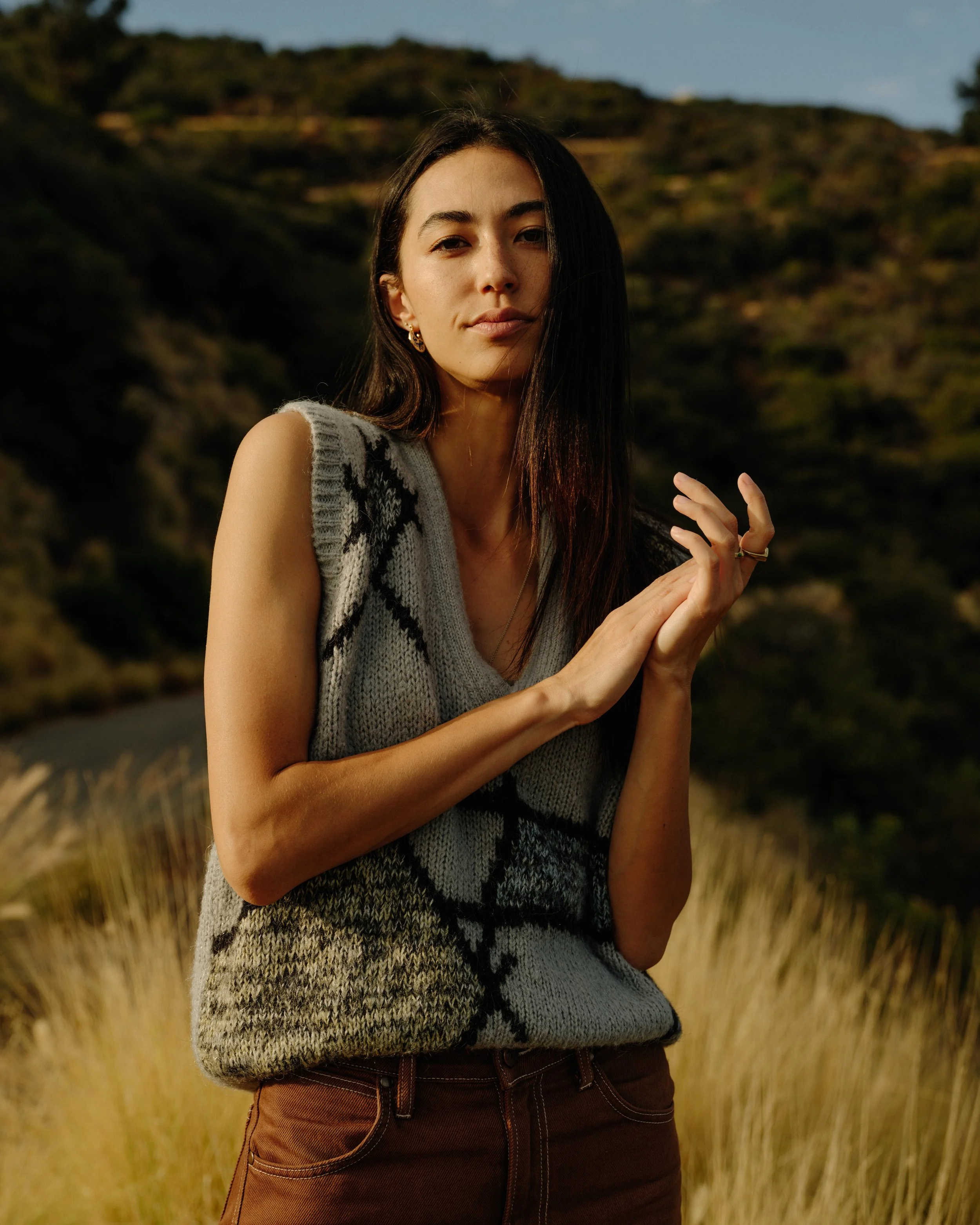 A young woman with long dark hair and earrings stands outdoors in a field of tall grass, with a rocky hillside in the background. She wears a sleeveless sweater with a black and gray pattern and brown pants, and looks directly at the camera.