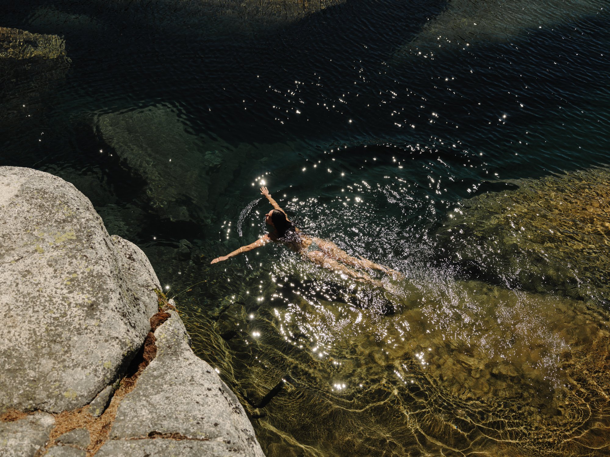 Person swimming in a natural body of water near rocks and sunlight reflecting on the water's surface.