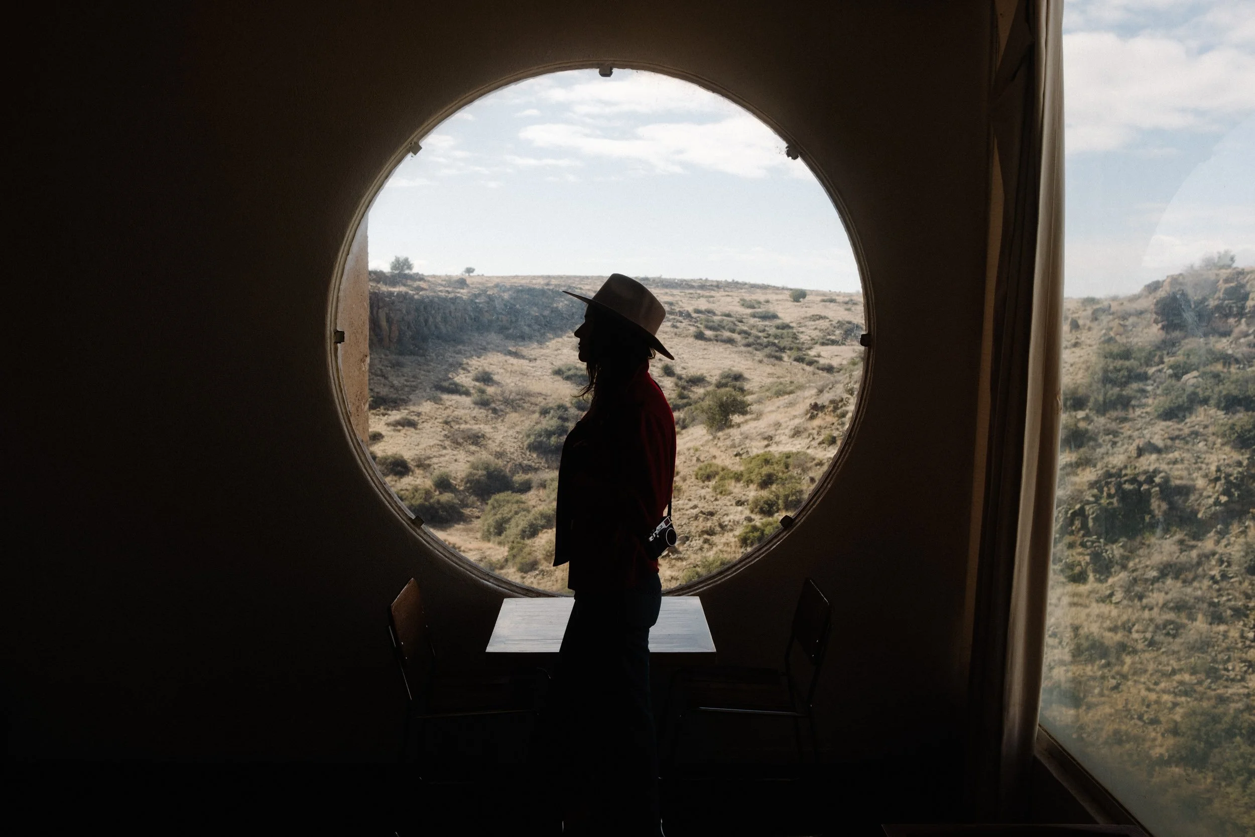 Silhouette of a person wearing a hat, standing in front of a large circular window with a desert landscape outside.